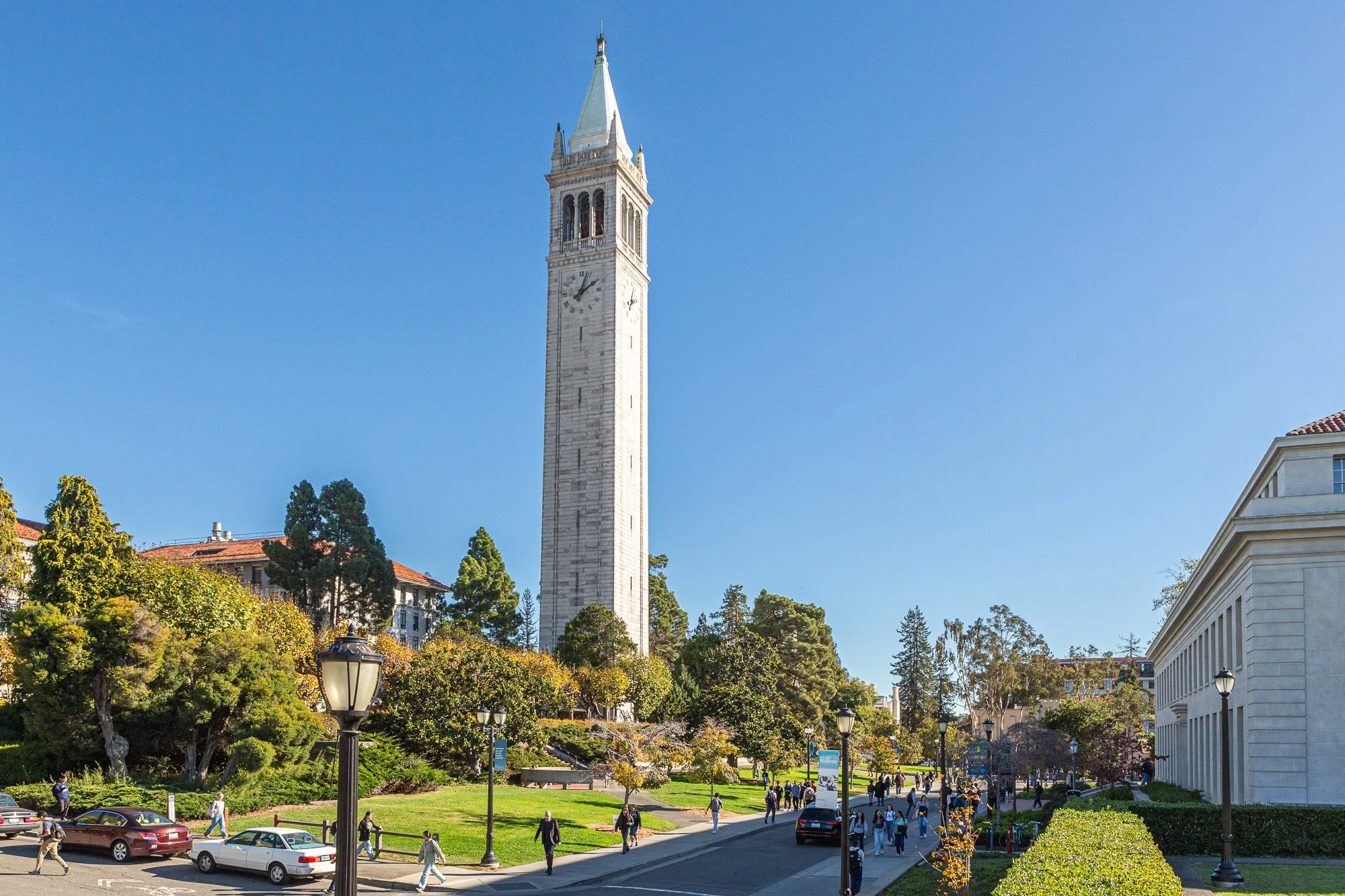 People walking on a sidewalk in front of a large clock tower surrounded by green trees and a white building, with a clear blue sky