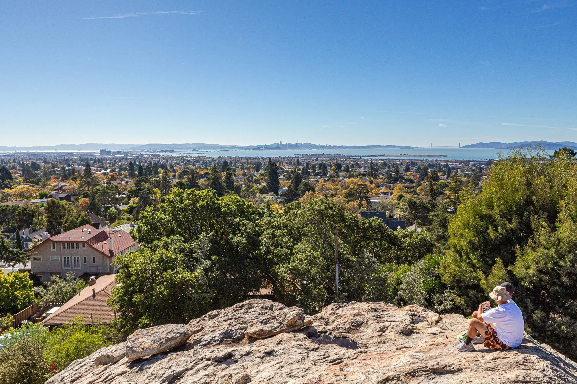 Person sitting on a rocky ledge overlooking a city and bay, with trees in the foreground and a clear blue sky above.