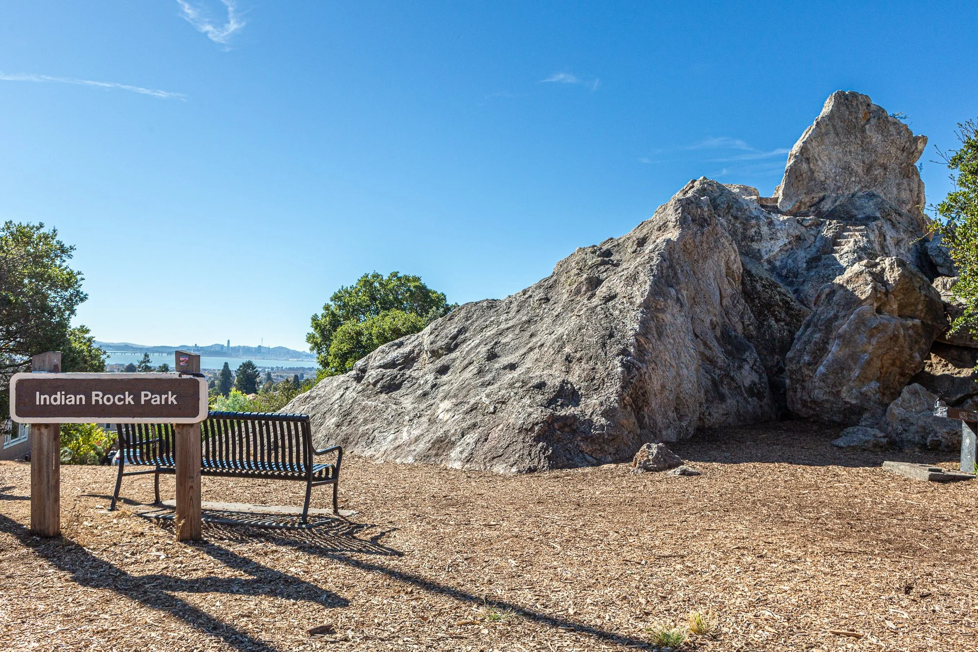 Large rocks at Indian Rock Park with a bench and a sign in the foreground, trees, city skyline, and blue sky in the background.