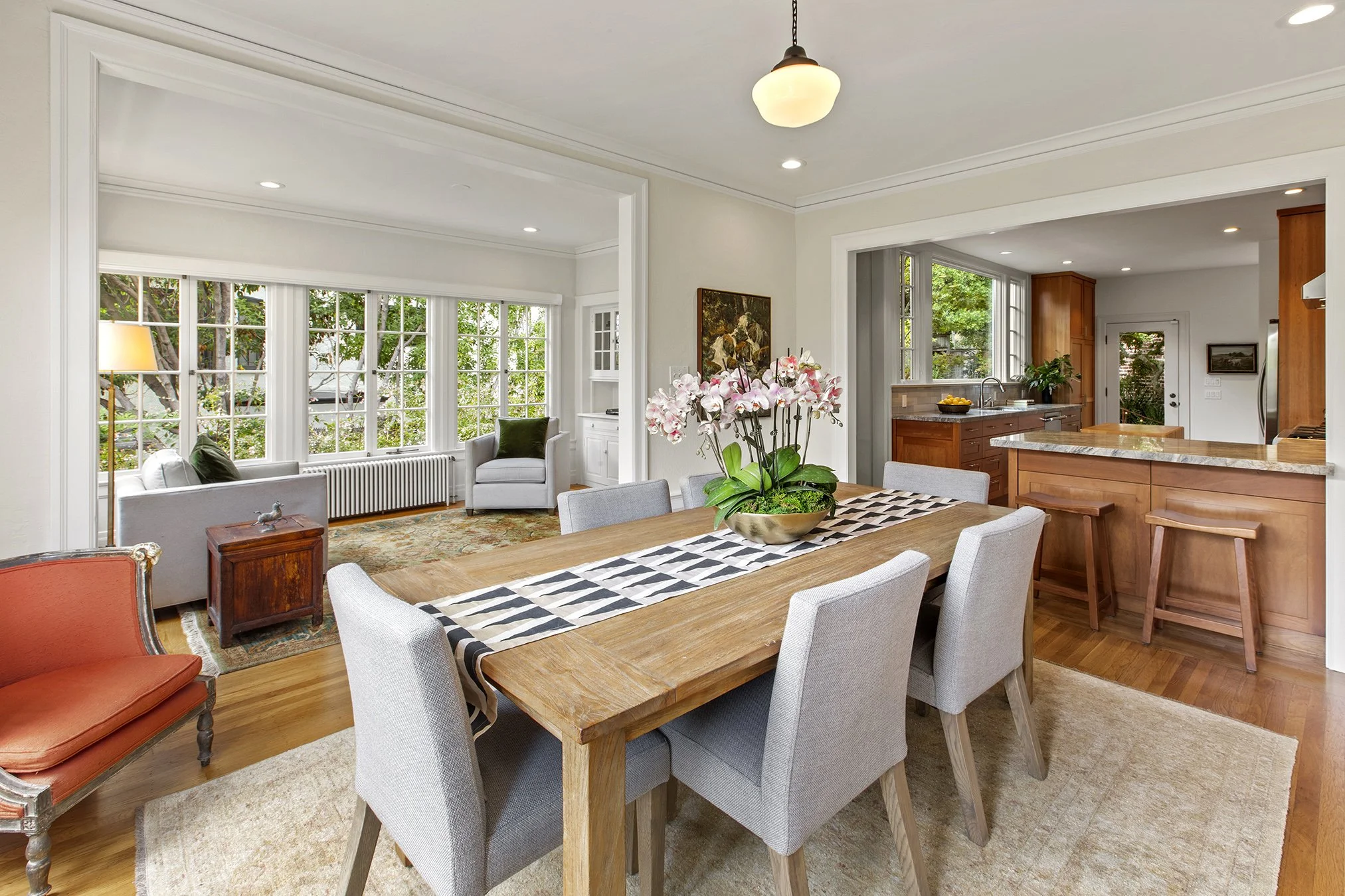 Open-concept living and dining area with large windows, a wooden dining table with a floral centerpiece, and a kitchen with wooden cabinets and a granite countertop.
