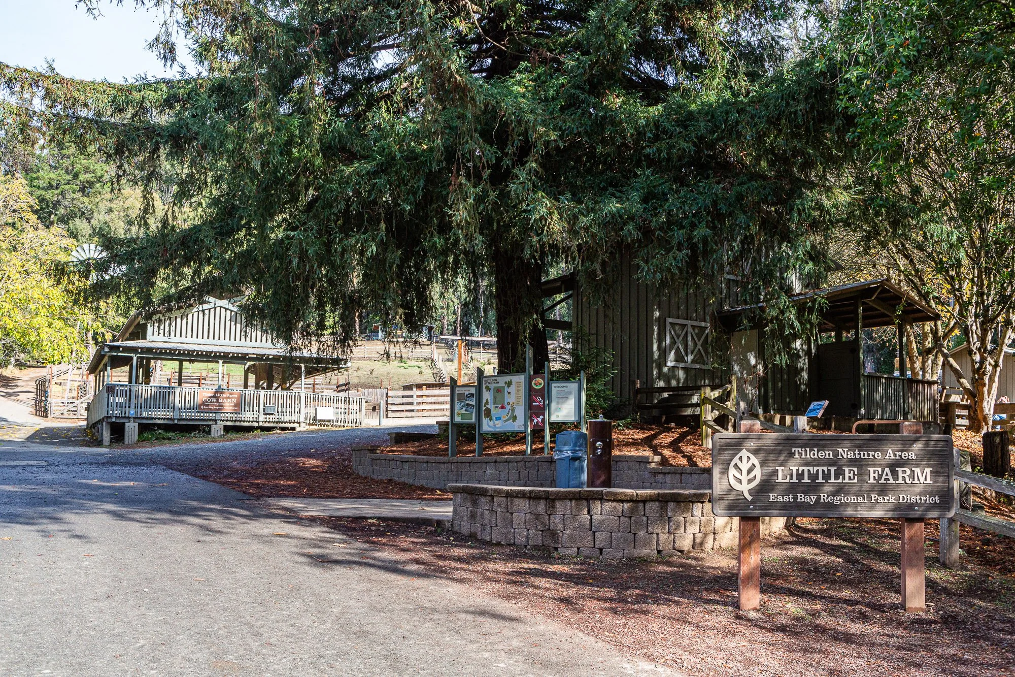 Entrance to Tilden Nature Area Little Farm with a sign and large trees, wooden barn structures, and informational boards in the background.
