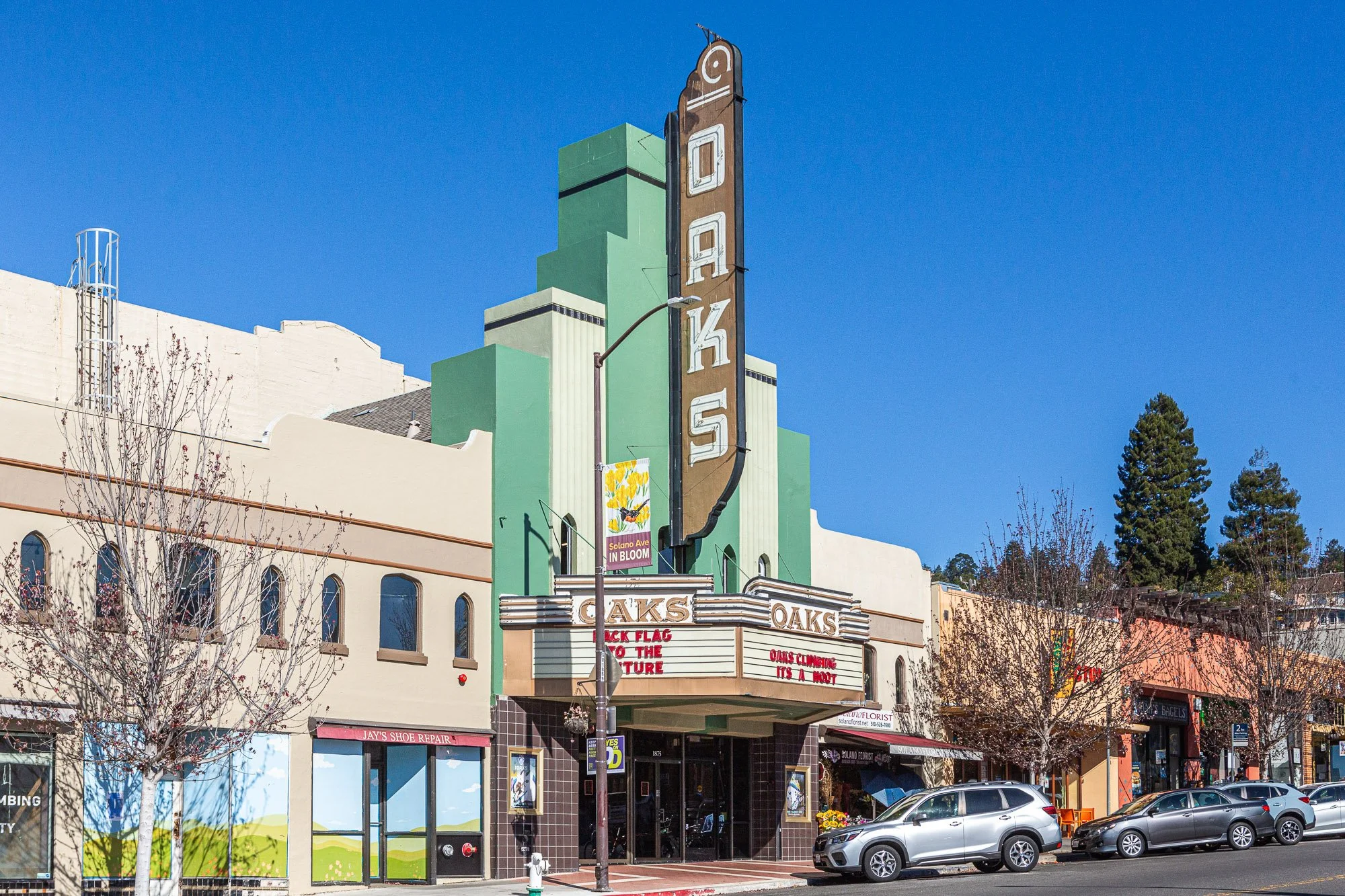 A historic theater named 'Oaks' with a large vertical marquee sign and an art deco architectural style, featuring colorful buildings and parked cars in the foreground.