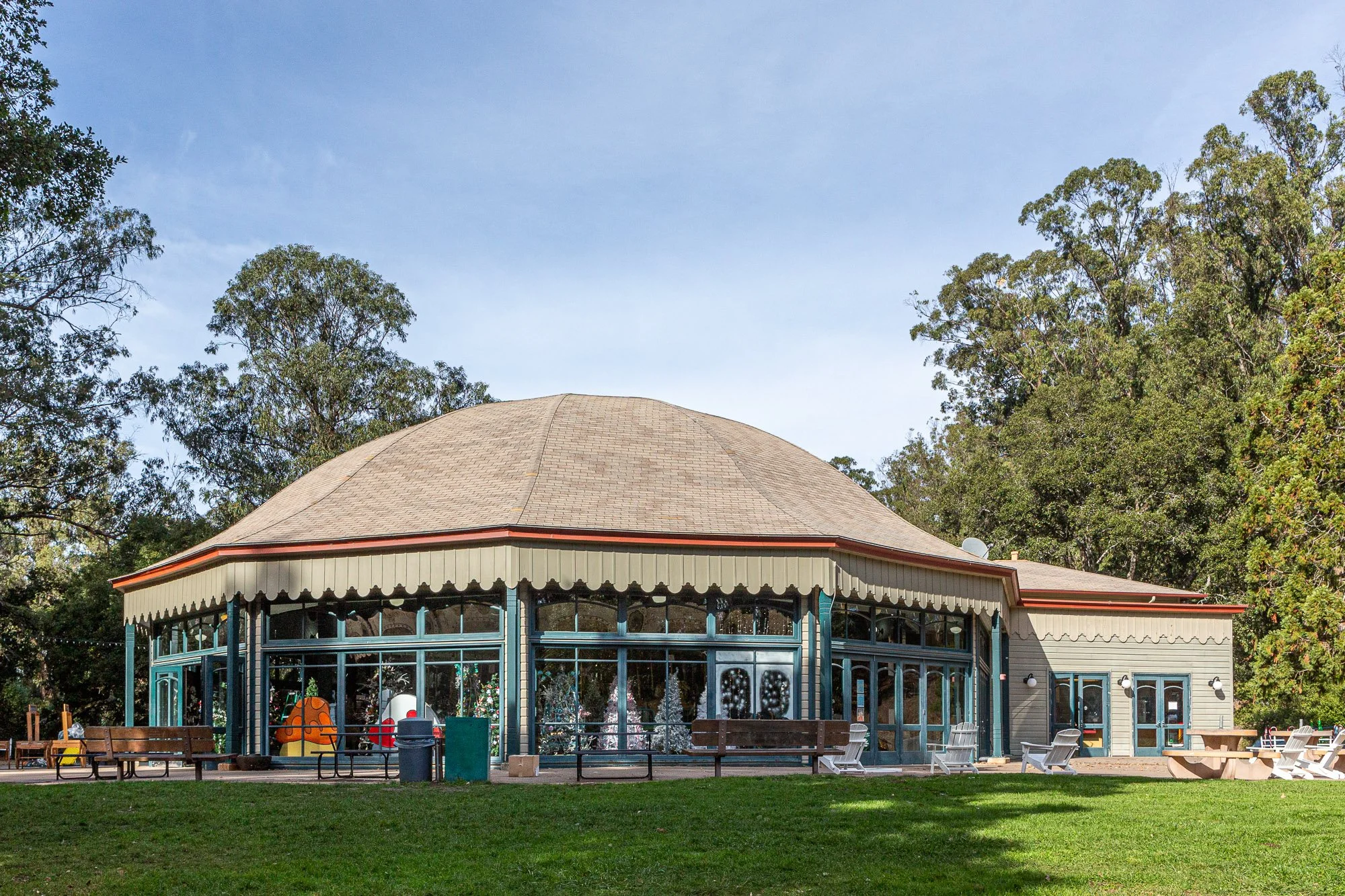 A round building with large glass windows, a beige shingled roof, and decorative trim, situated in a park with green grass and trees.