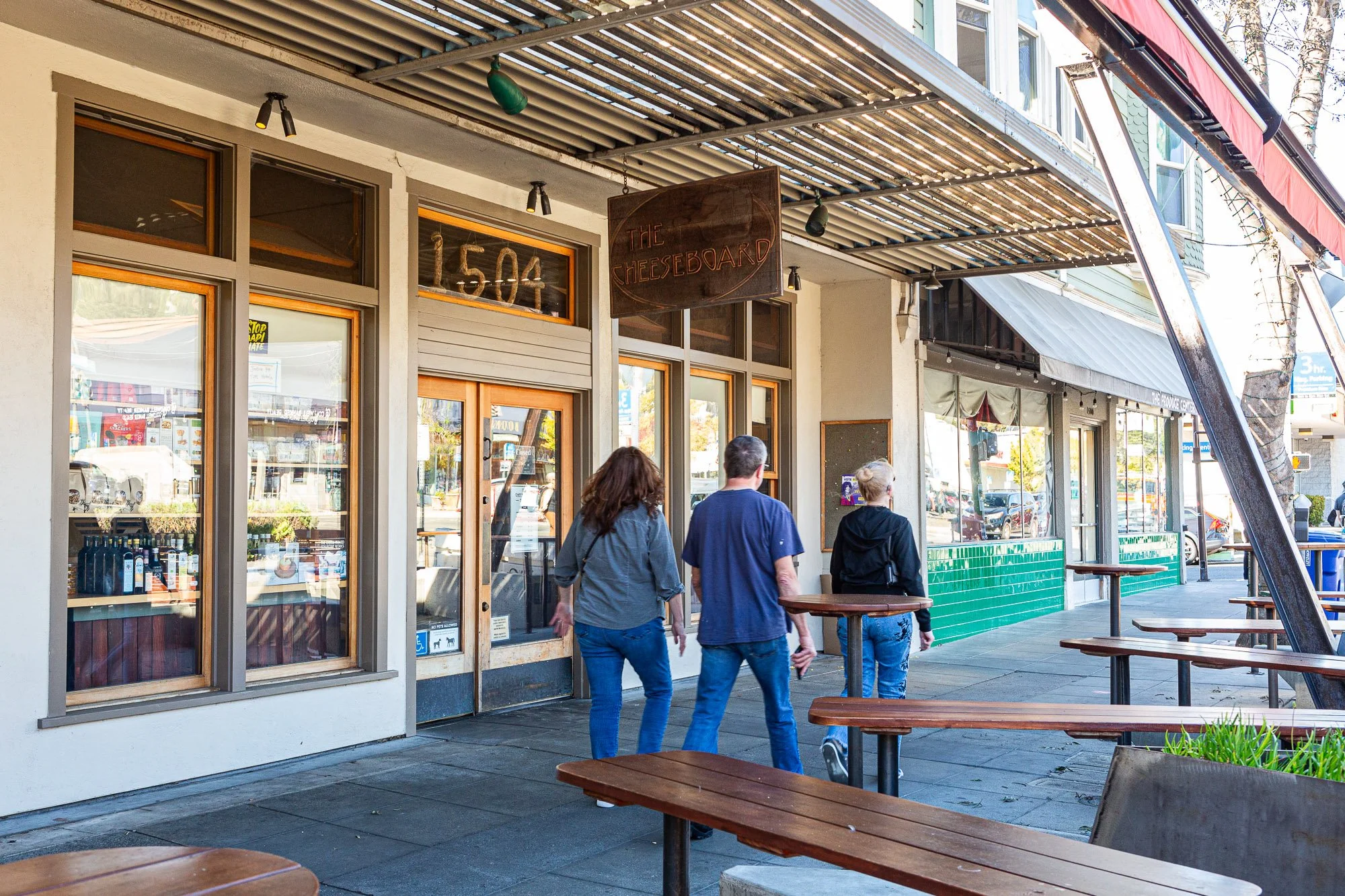 People walking outside a storefront called The Cheeseboard, with outdoor seating and green tiled wall, on a sunny day.