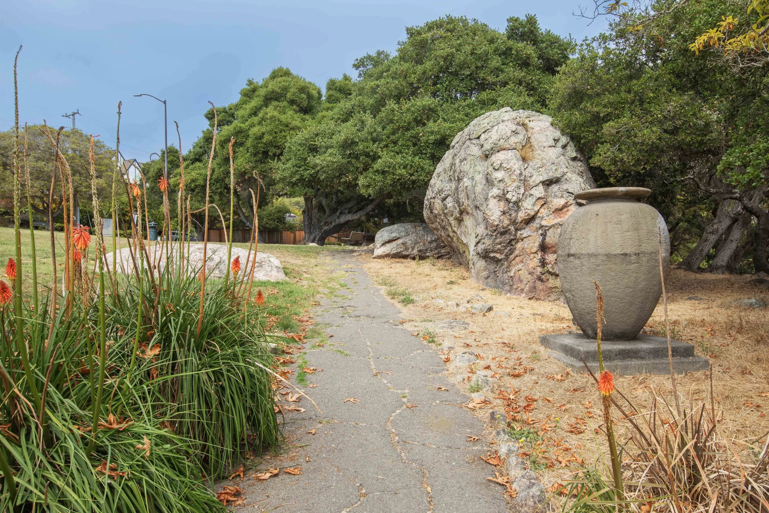 A park pathway with large rocks, a stone urn, green bushes, and trees with overcast sky.
