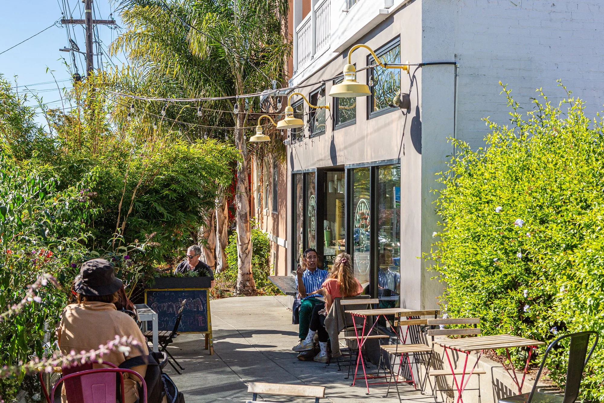 People sitting outside at a cafe on a sunny day, surrounded by lush greenery and tropical trees, with hanging string lights overhead.