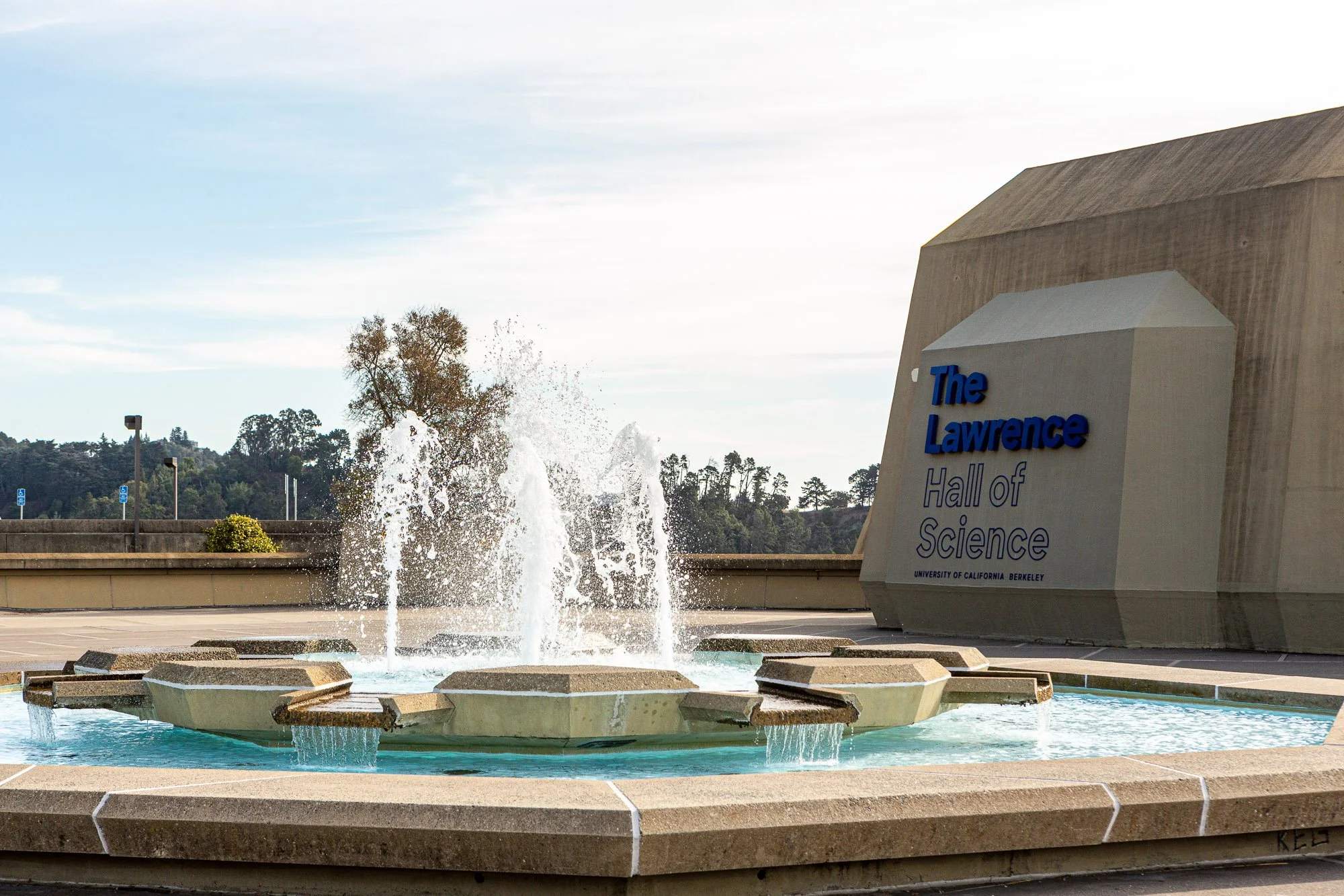 The entrance to the Lawrence Hall of Science at the University of California, Berkeley, featuring a fountain with waterfalls in the foreground and a large modern building with the hall's name on it.