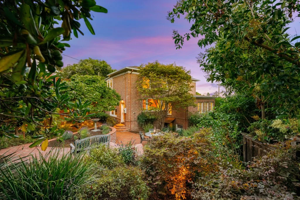 A brick house surrounded by lush green trees and plants under a colorful twilight sky.