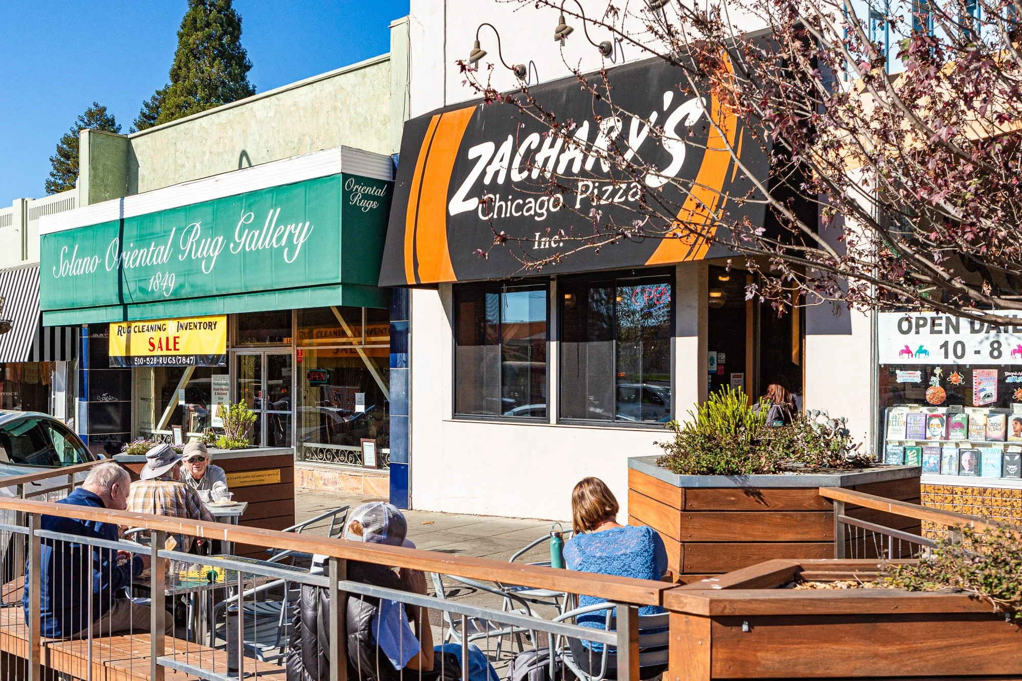 People sitting outside cafes on a sunny day, with storefronts including a rug gallery and a restaurant named Zachary's Chicago Pizza, and signage indicating opening hours and sales.