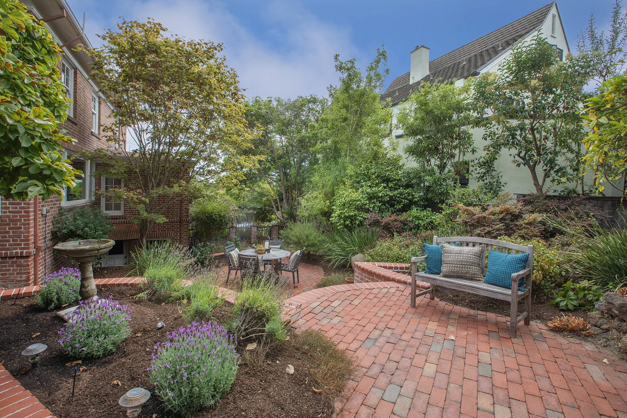 Garden backyard with brick paving, a wooden bench with cushions, a round table with chairs, lush green trees and shrubs, and a birdbath, under a partly cloudy sky.