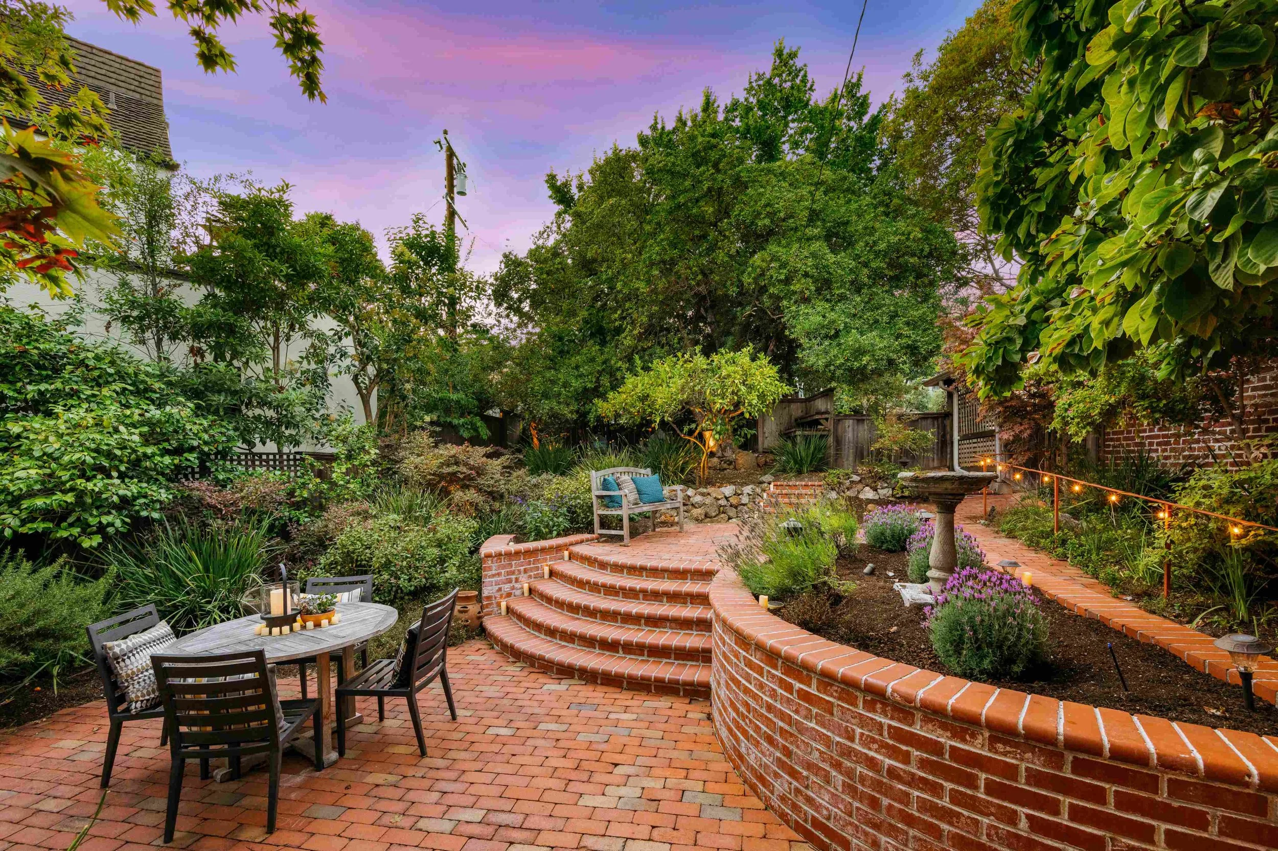 A backyard garden with a brick patio, seating area, lush green trees, flowering plants, a bird bath, and string lights, during sunset.