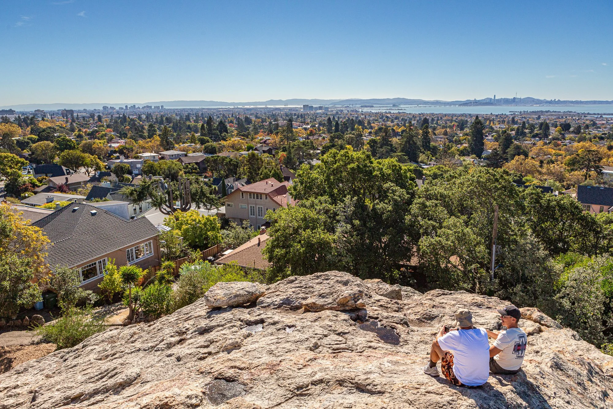 Two people sitting on a large rock, overlooking a cityscape with houses, trees, and a distant view of water and skyline on a sunny day.