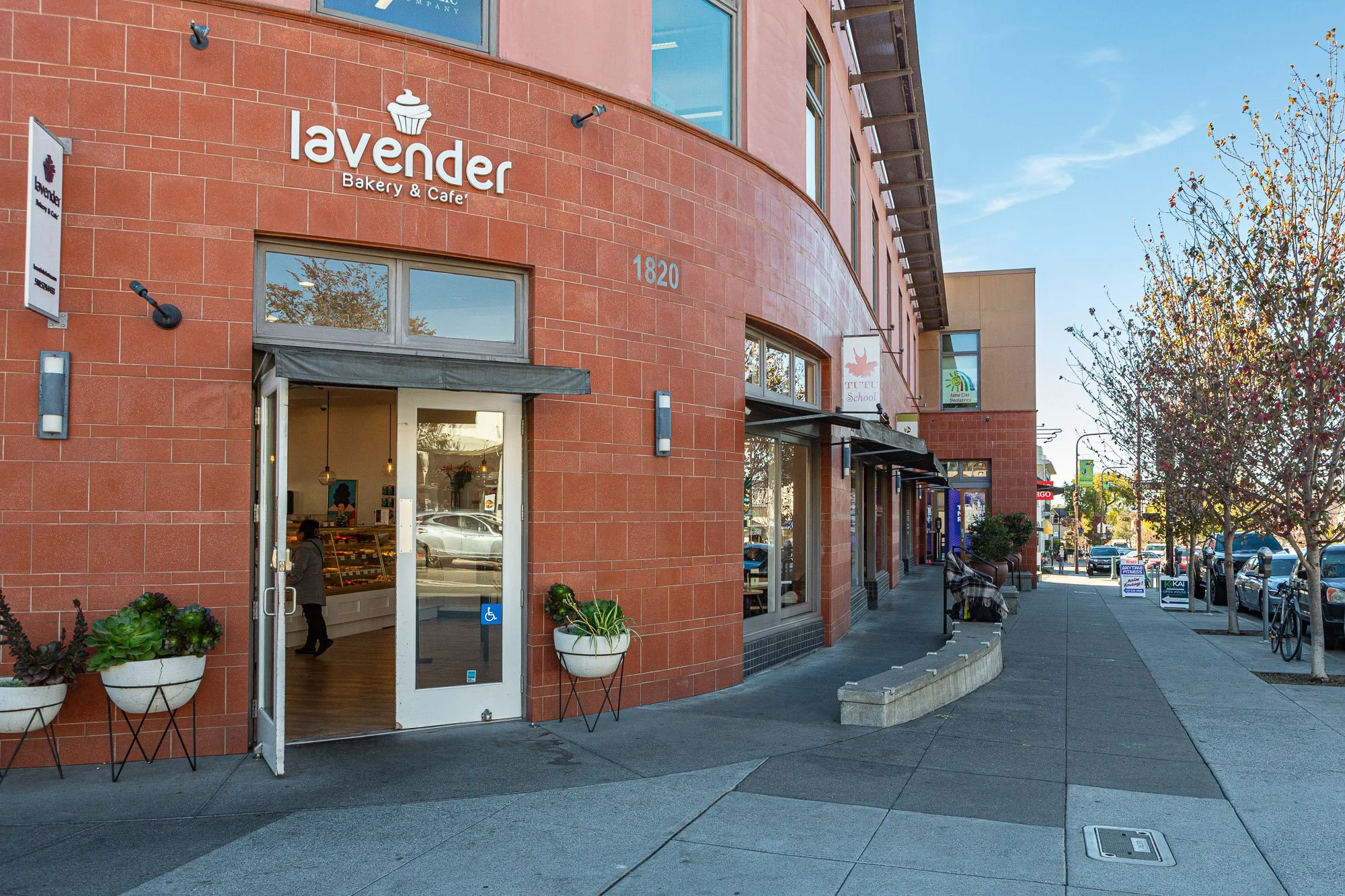 Exterior view of Lavenda Bakery & Cafe on a sunny day, showing a brick building with sidewalk seating, potted plants outside, and trees lining the street.
