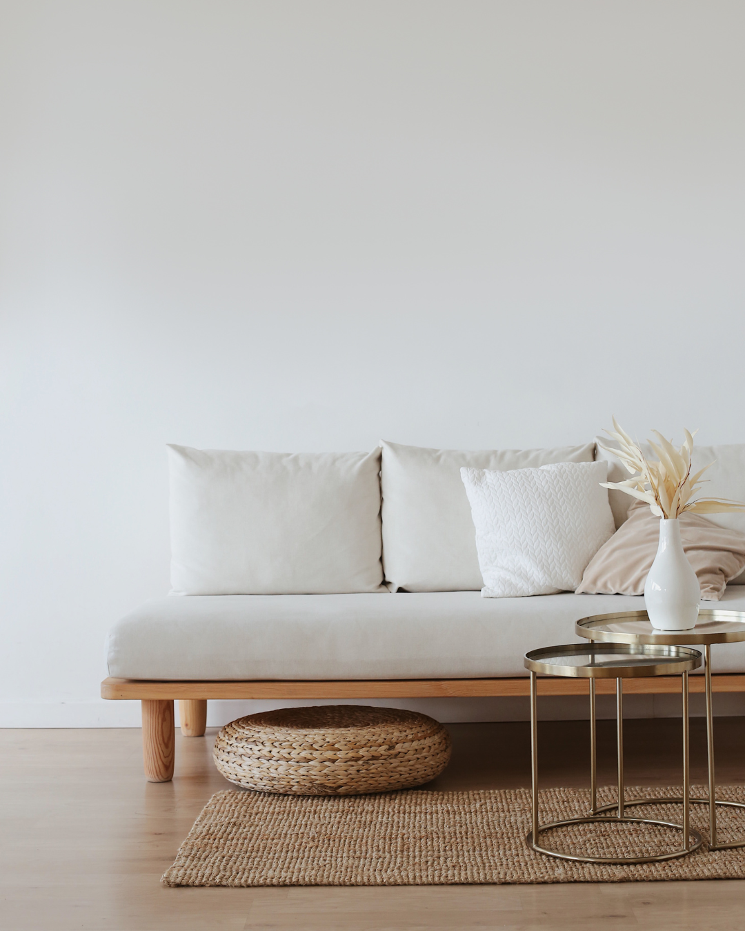 Minimalist living room with a beige sofa, white pillows, wicker ottoman, and two nested round metal trays with a white vase and dried leaves on a small table, on a natural fiber rug.