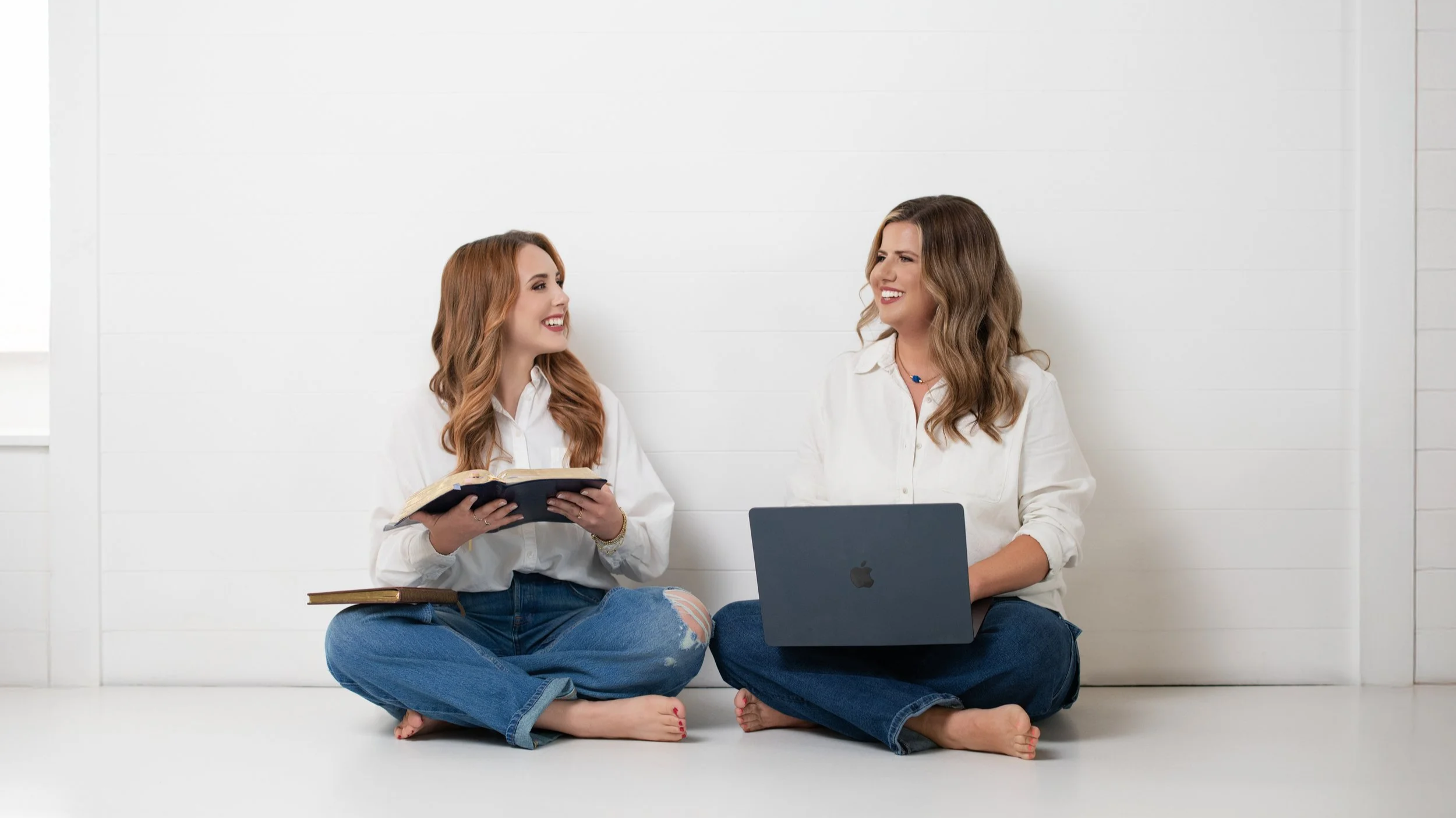 Owners of Launched Studios.  Two women sitting on the floor against a white wall, smiling at each other. One is holding an open book, and the other has a laptop on her lap, both wearing white shirts and blue jeans.