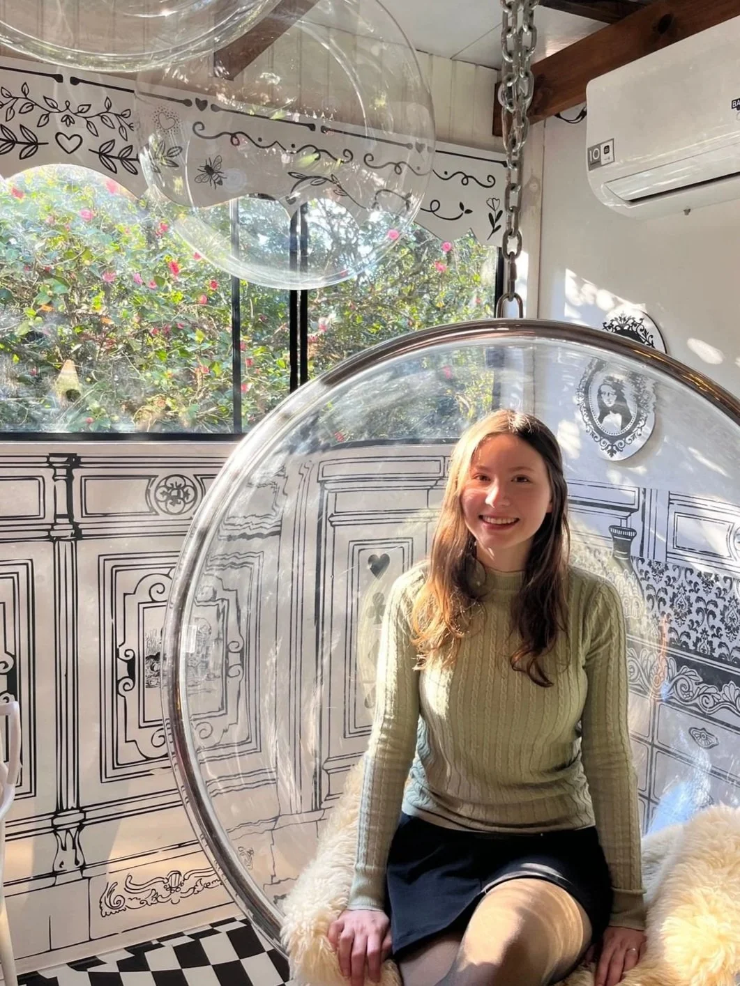 Young woman with long hair smiling while sitting inside a transparent hanging bubble chair in a decorated room with black and white wall art and a window showing trees and pink flowers outside.