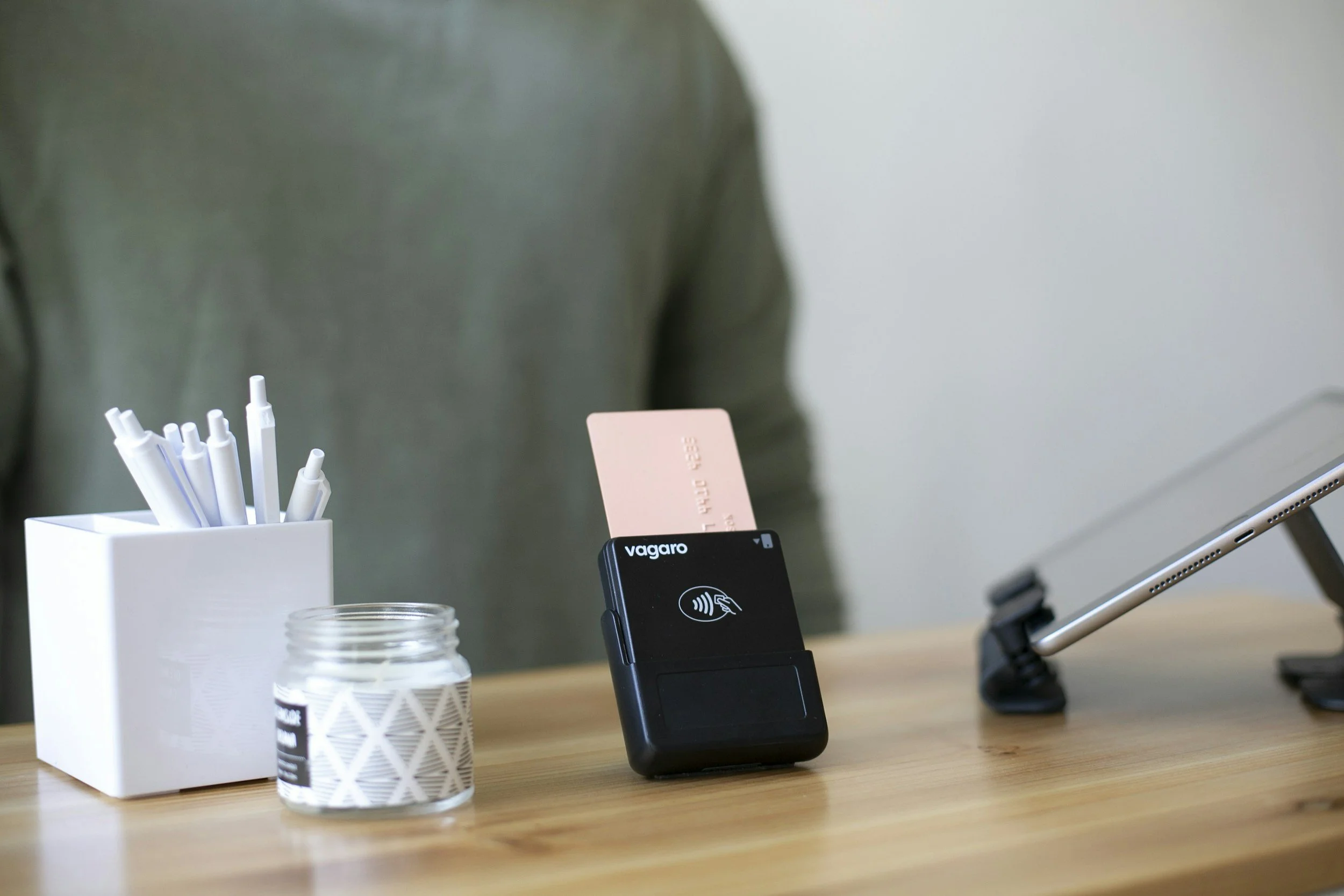 A wooden desk with a white pen holder containing pens, a white decorative candle, a black contactless payment device with a pink card inserted, and a tablet on a stand.