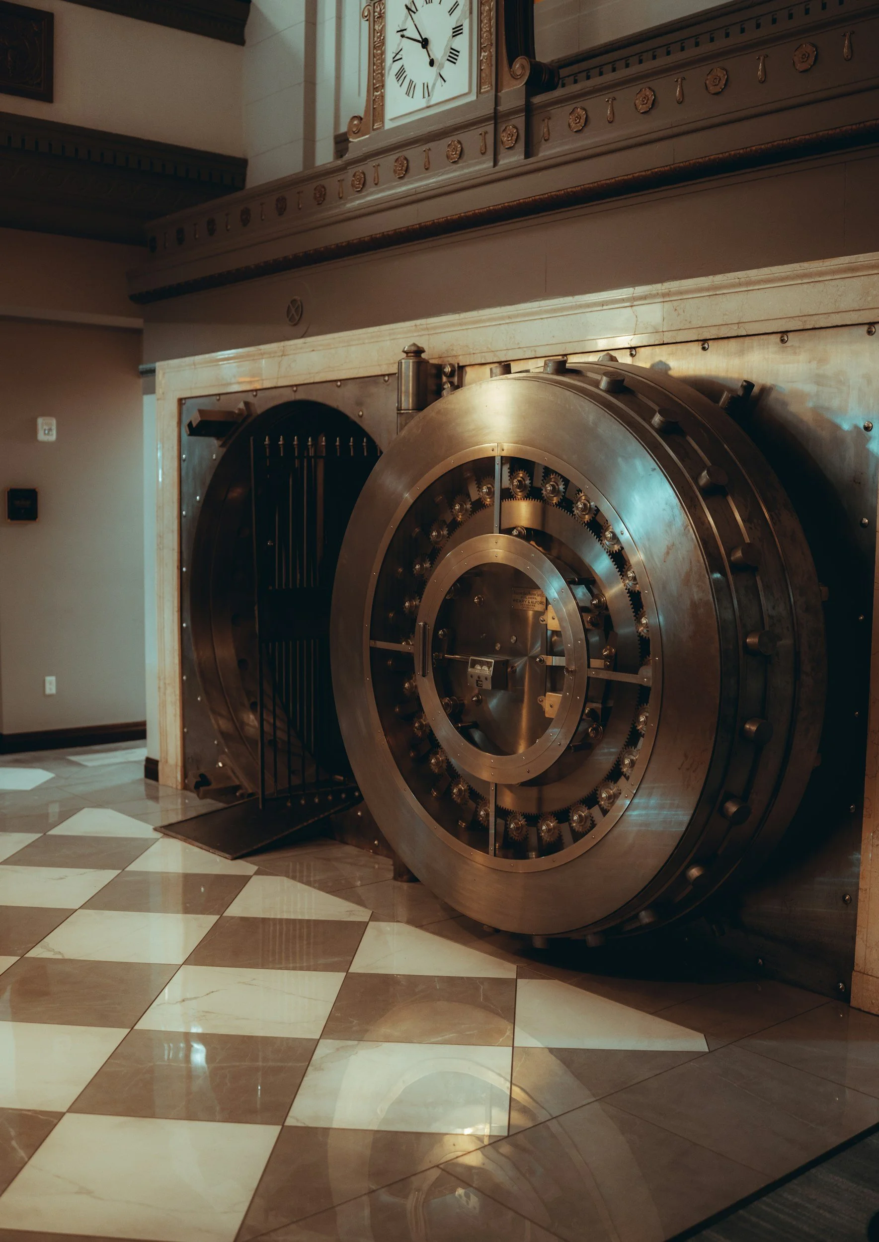 Large banking vault door with a combination dial, open to reveal the dark interior, situated in a lobby with checkered floor tiles and a decorative clock above.