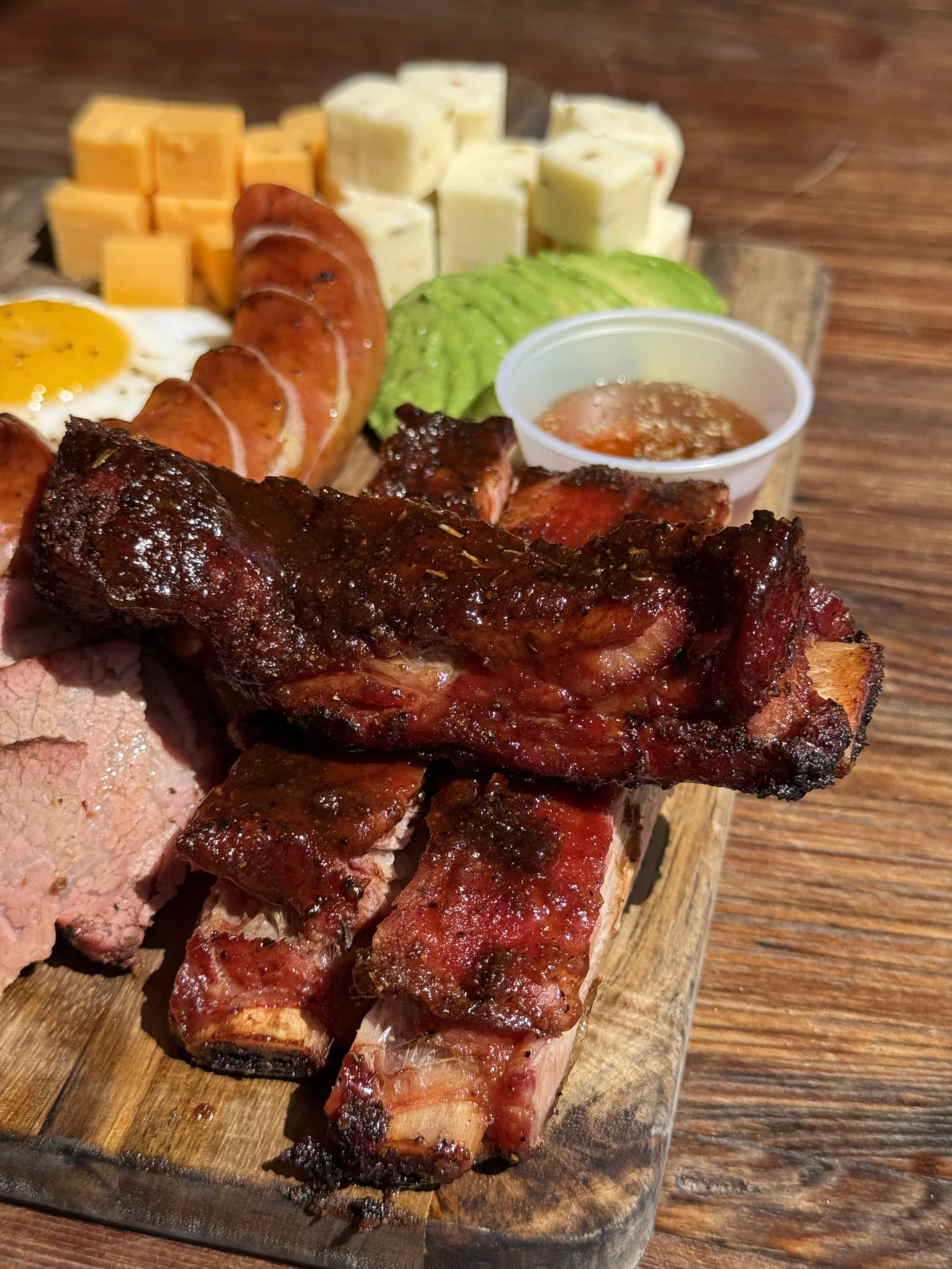 A wooden platter with grilled meats, cheese cubes, sliced sausage, half a boiled egg, avocado slices, and a cup of barbecue sauce.