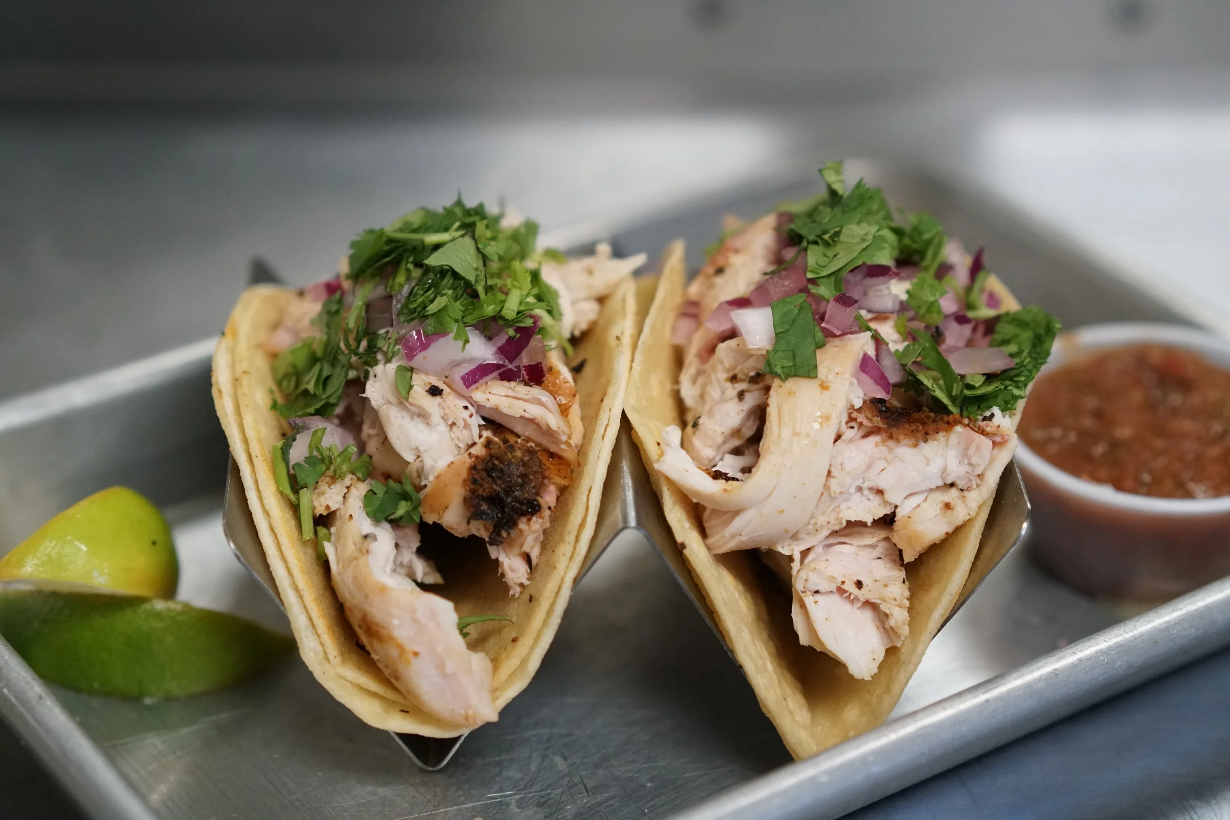 Chicken tacos with chopped onions and cilantro, served with lime wedges and salsa on a metal tray.