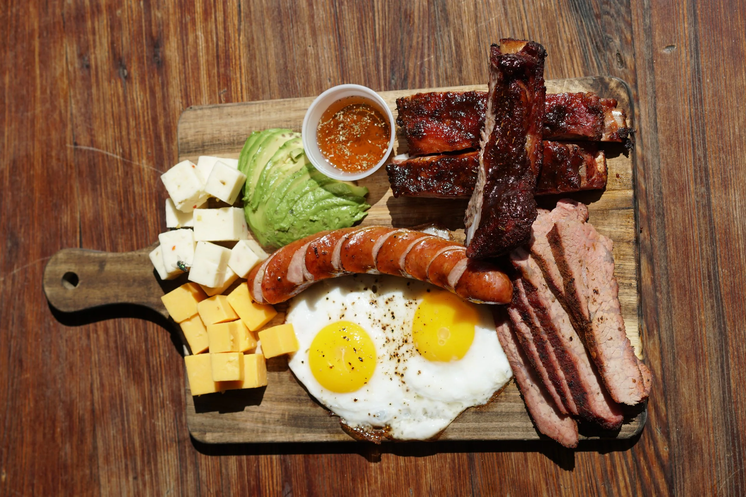 Breakfast platter with fried eggs, bacon, sausage, ribs, cheese, avocado slices, cheese cubes, and a small cup of syrup or honey on a wooden serving board.