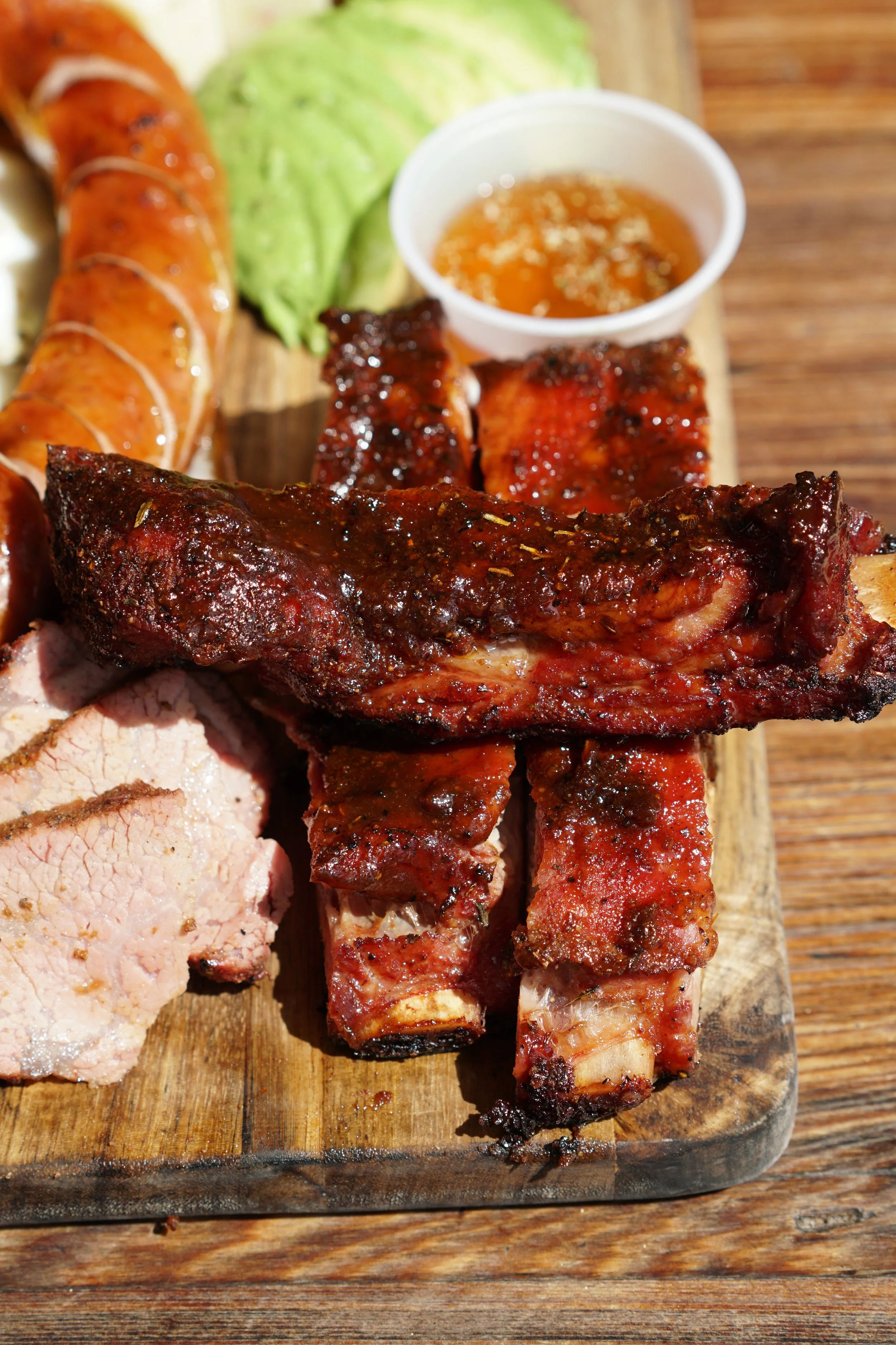 Close-up of a wooden platter with grilled meats including ribs and sausage, accompanied by a small cup of barbecue sauce, lettuce, and sliced roast meat, on a wooden table.