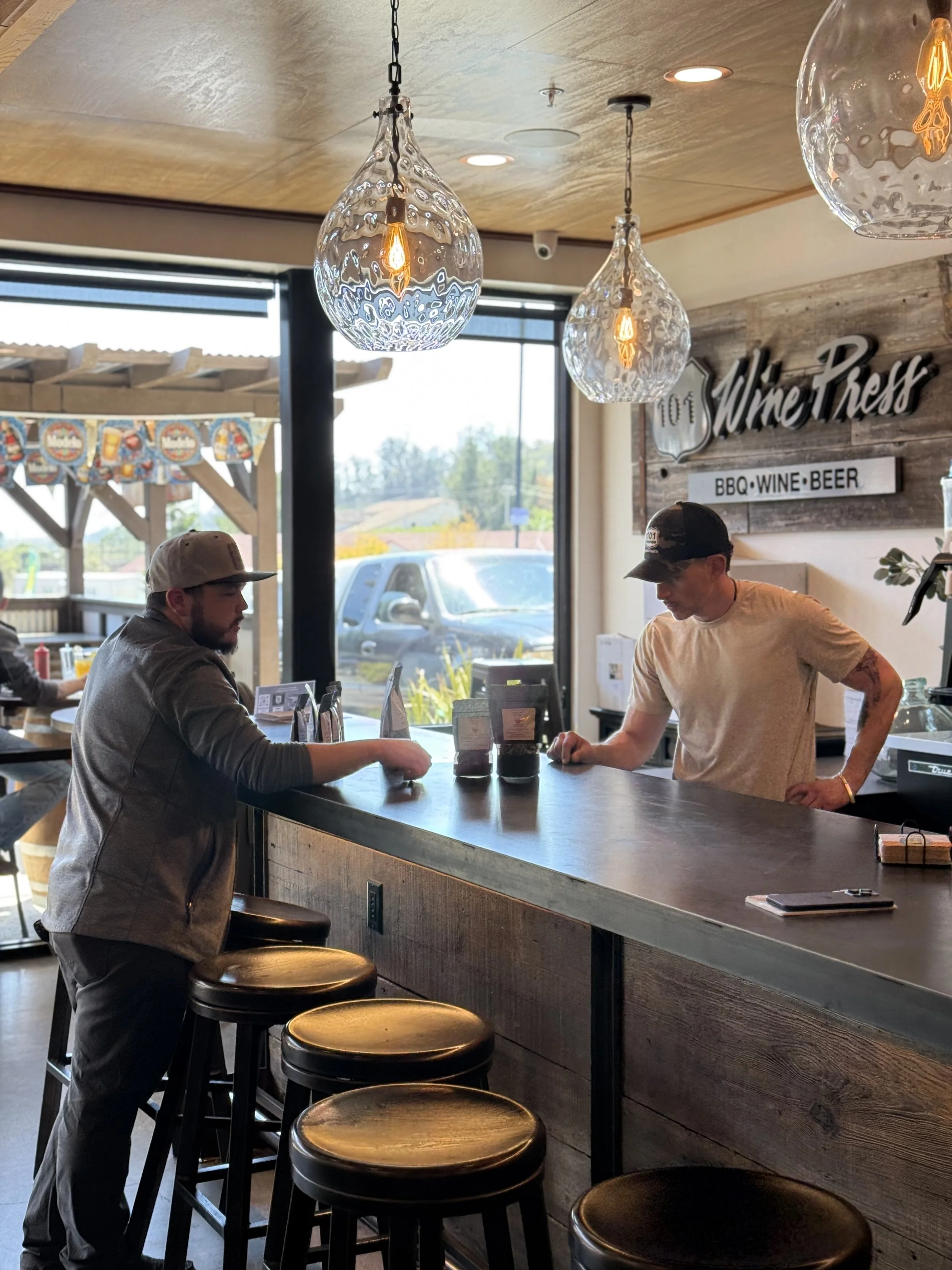 Inside a restaurant named 'Wine Press' with a bar counter, two men standing and conversing, hanging glass light fixtures, large window showing parked cars outside, and a sign indicating they serve BBQ, wine, and beer.