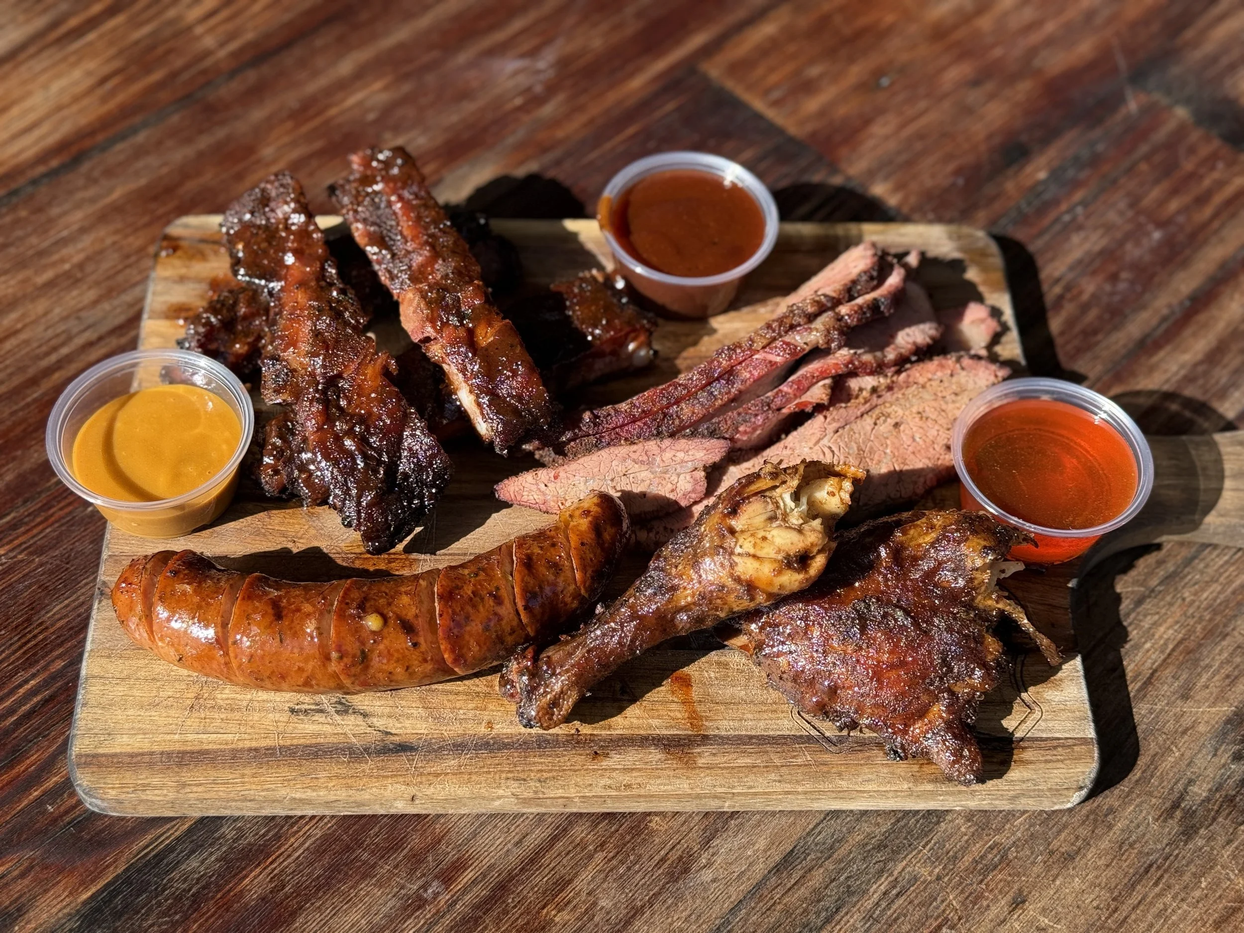 Assorted barbecue meats with three small cups of barbecue sauce on a wooden serving board.