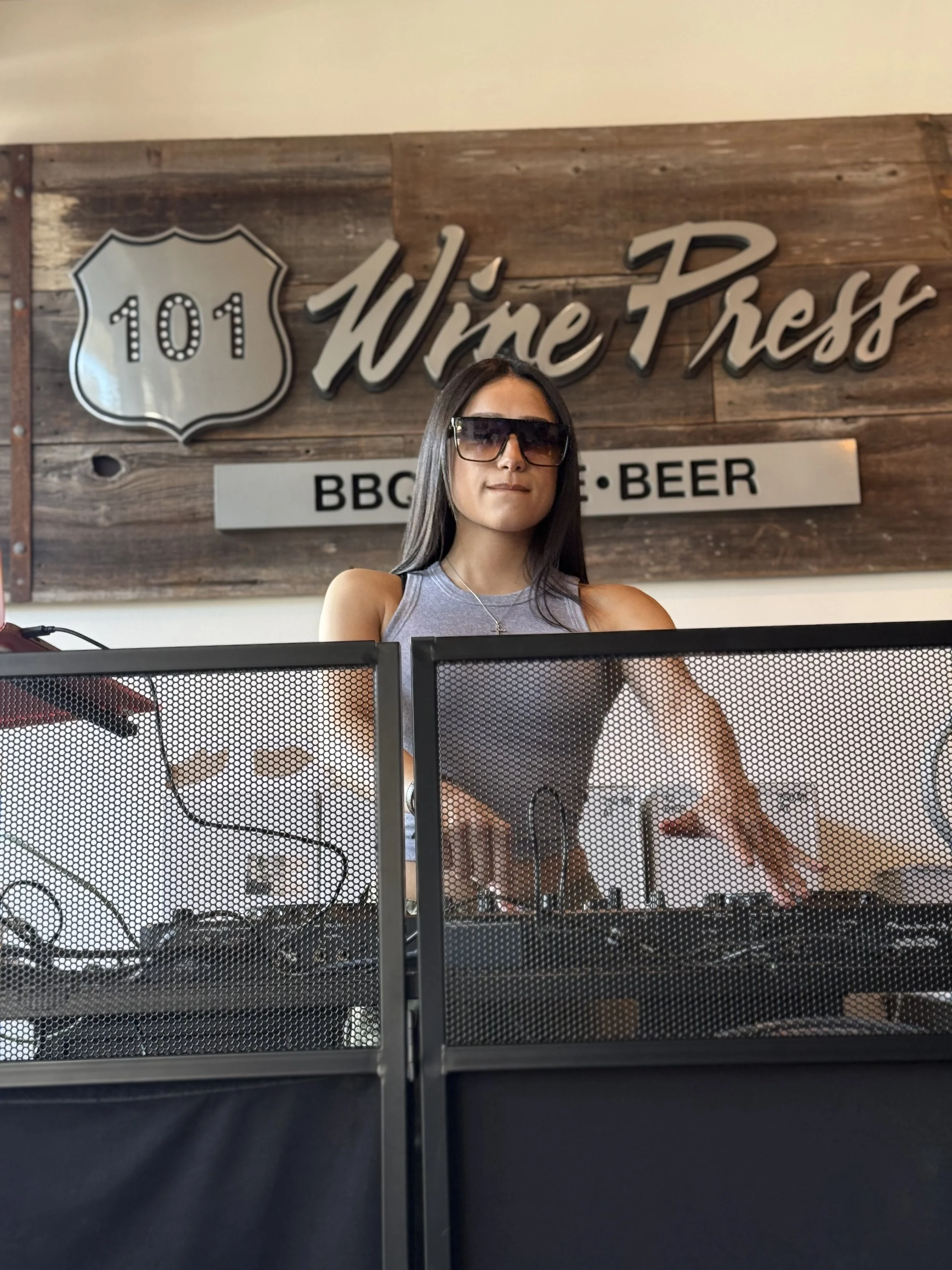 A woman DJ wearing large sunglasses standing behind DJ equipment at a restaurant called Wire Fresh, with a sign behind her reading 'BBQ, Beer'.