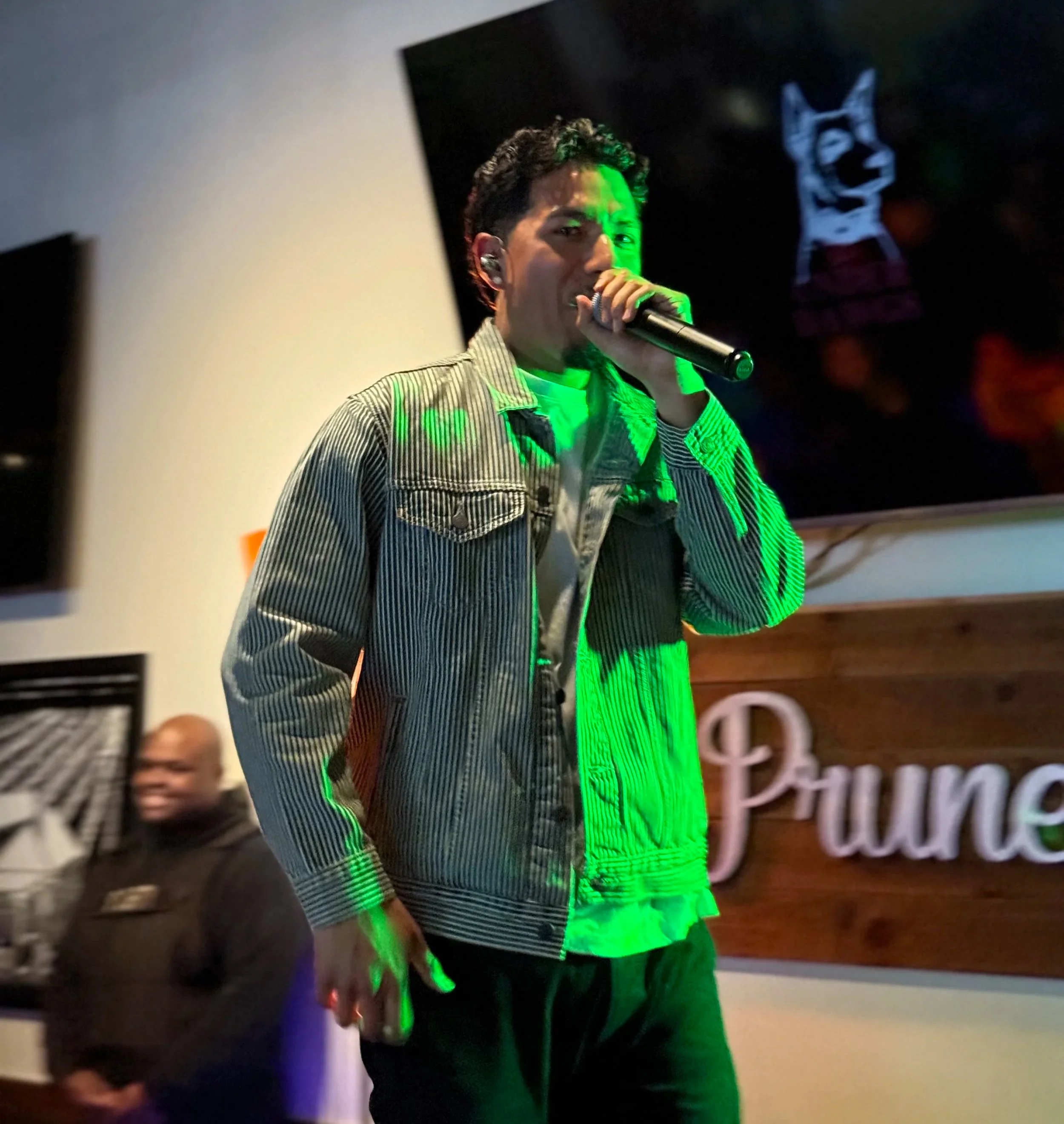 A young man singing into a microphone on stage, wearing a striped jacket and green lighting illuminating him, with a poster and a man in a uniform in the background.