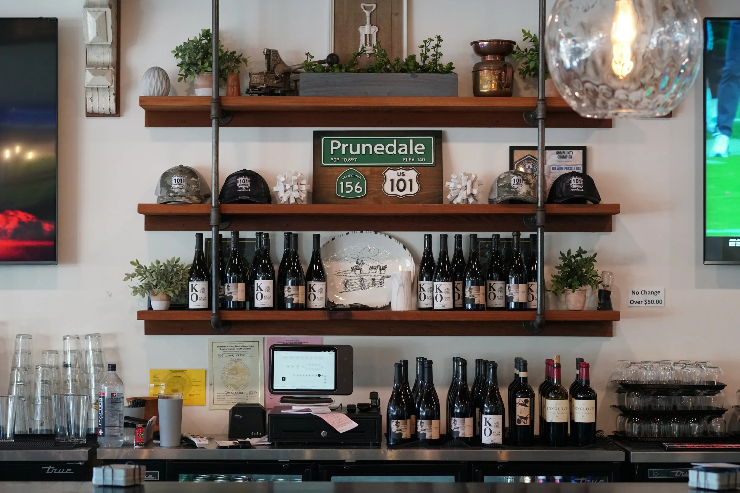 Wall display for the 101 Wine Press BBQ in Prunedale California. A bar with shelves holding liquor bottles, wine bottles, decorative items, helmets, and signs, with a counter below containing glasses, a cash register, and related items.