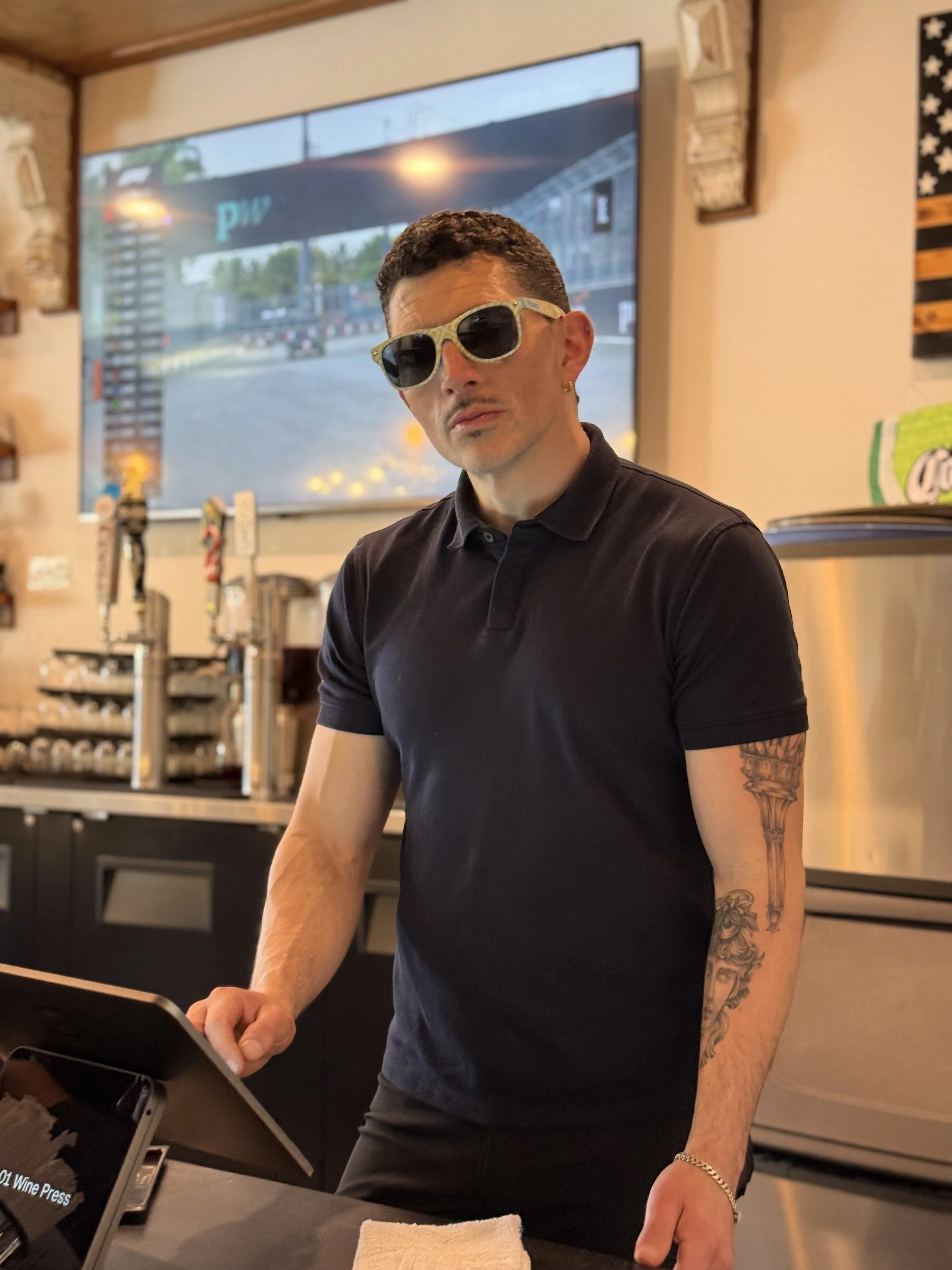 A man with dark hair, sunglasses, a black polo shirt, tattoos on his left arm, standing behind a counter in a bar or restaurant, with a large screen behind him showing a racing event.