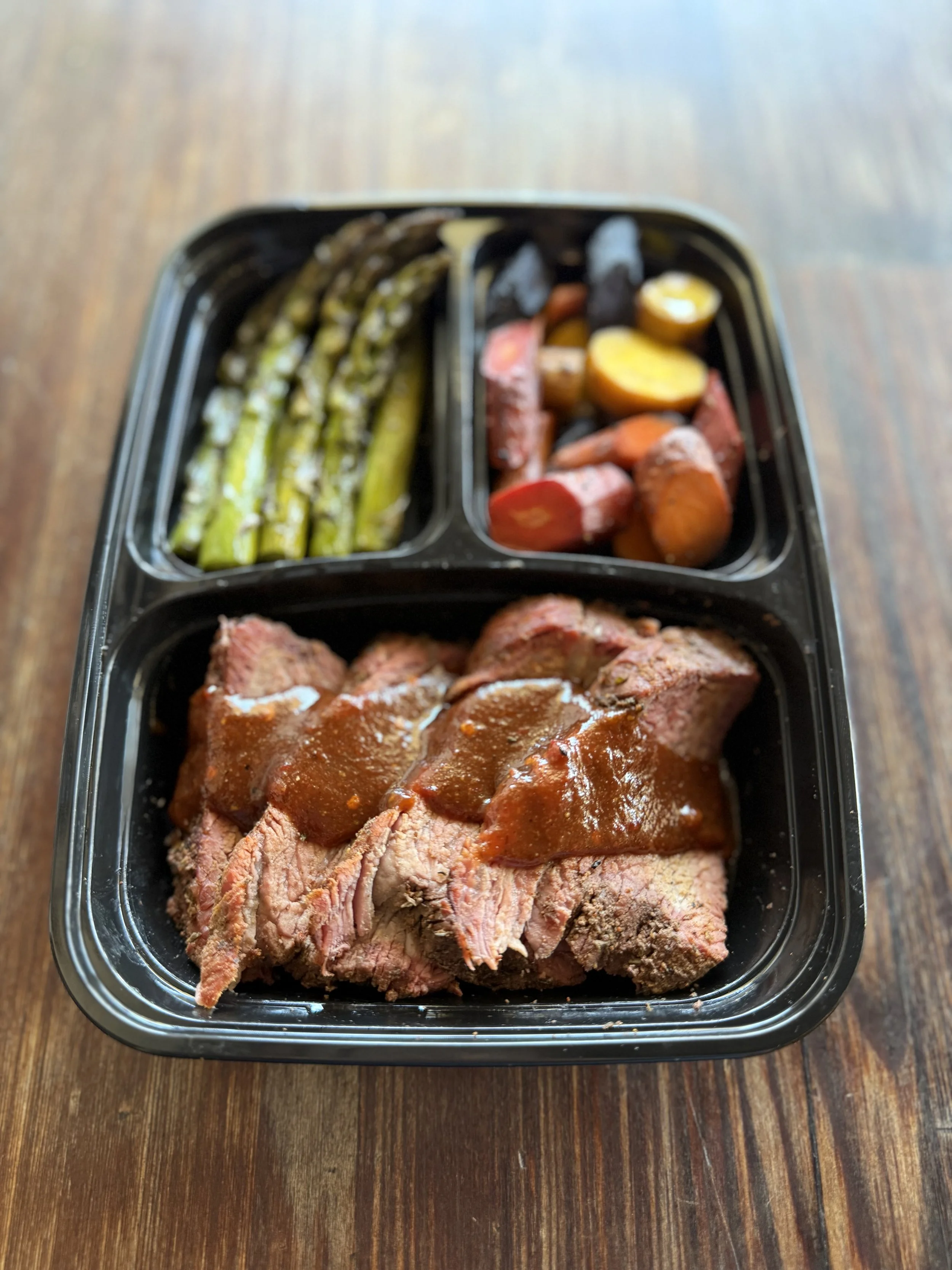 A meal in a divided black plastic meal prep container featuring sliced cooked roast beef with gravy, roasted carrots and sweet potatoes, and grilled asparagus.
