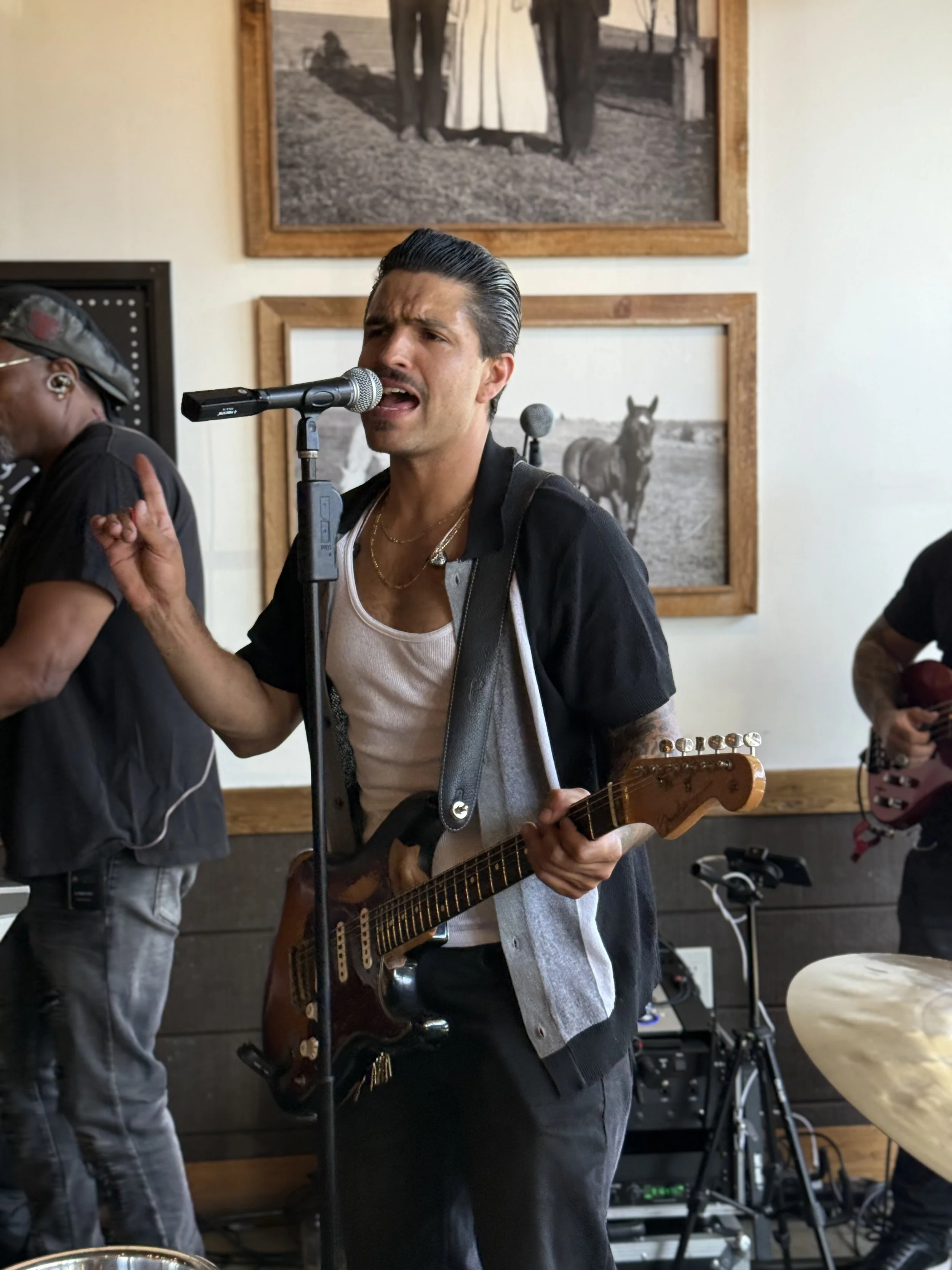 A man singing into a microphone while playing an electric guitar during a live performance. Another musician is partially visible playing an instrument on the right, and a person on the left is adjusting equipment. Behind them, there are framed black-and-white photographs on the wall depicting horses and a person in a white dress.