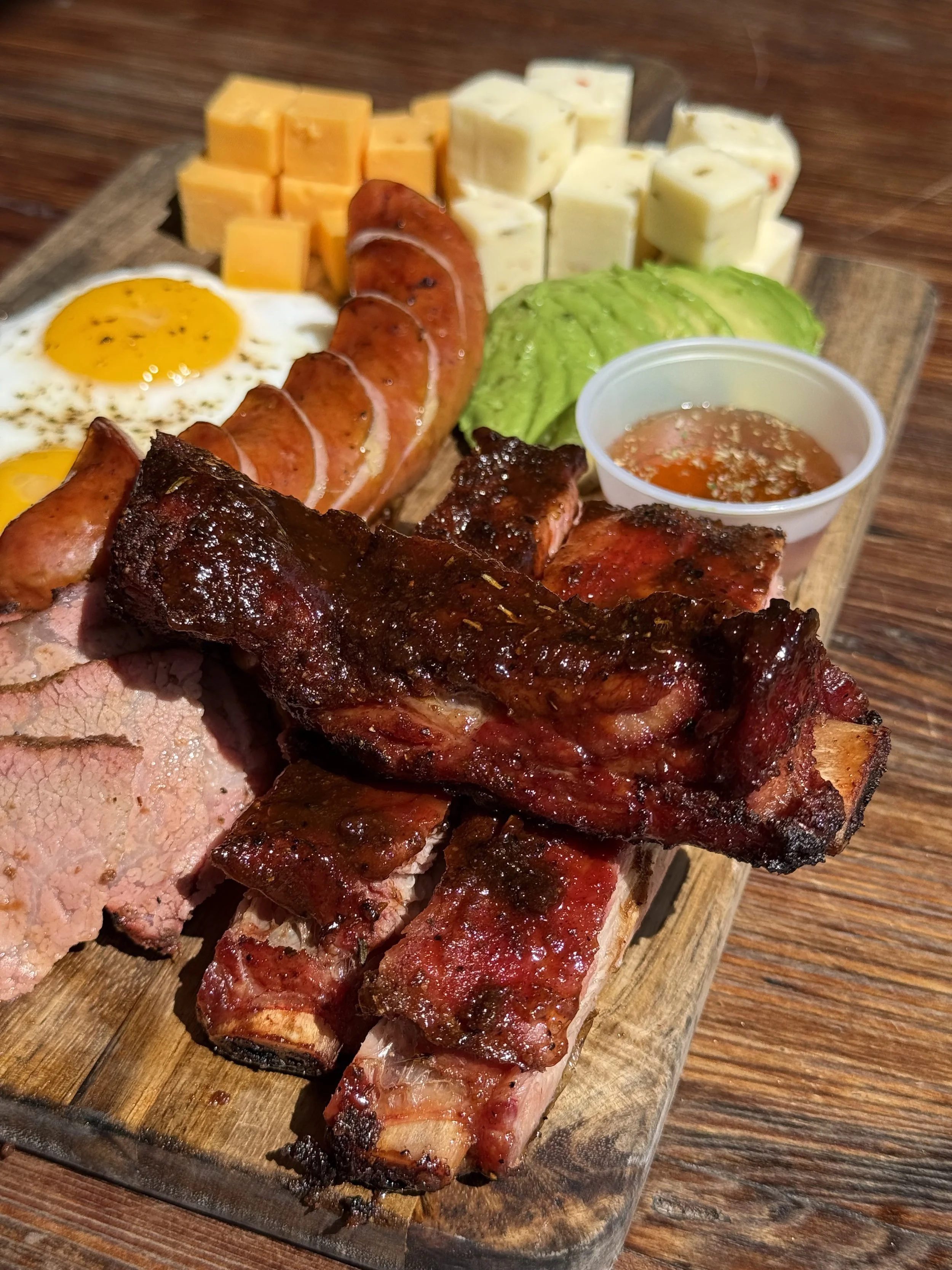 A wooden serving board with eggs, cheese, sausage, turkey, ribs, lettuce, and a small cup of sauce.