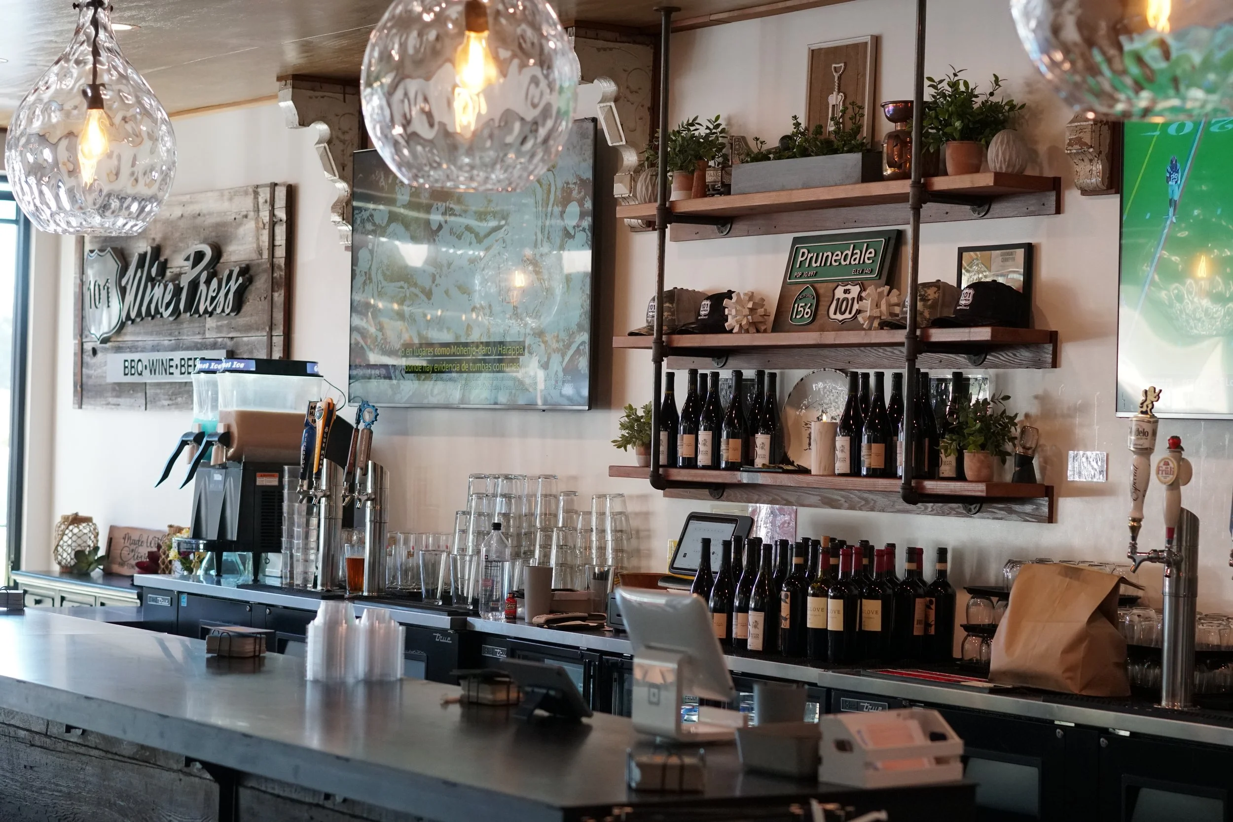 101 Wine Press BBQ bar with shelves holding wine bottles, potted plants, and decorative signs, a countertop with glasses and a tablet, and hanging glass light fixtures in a rustic decorated setting.