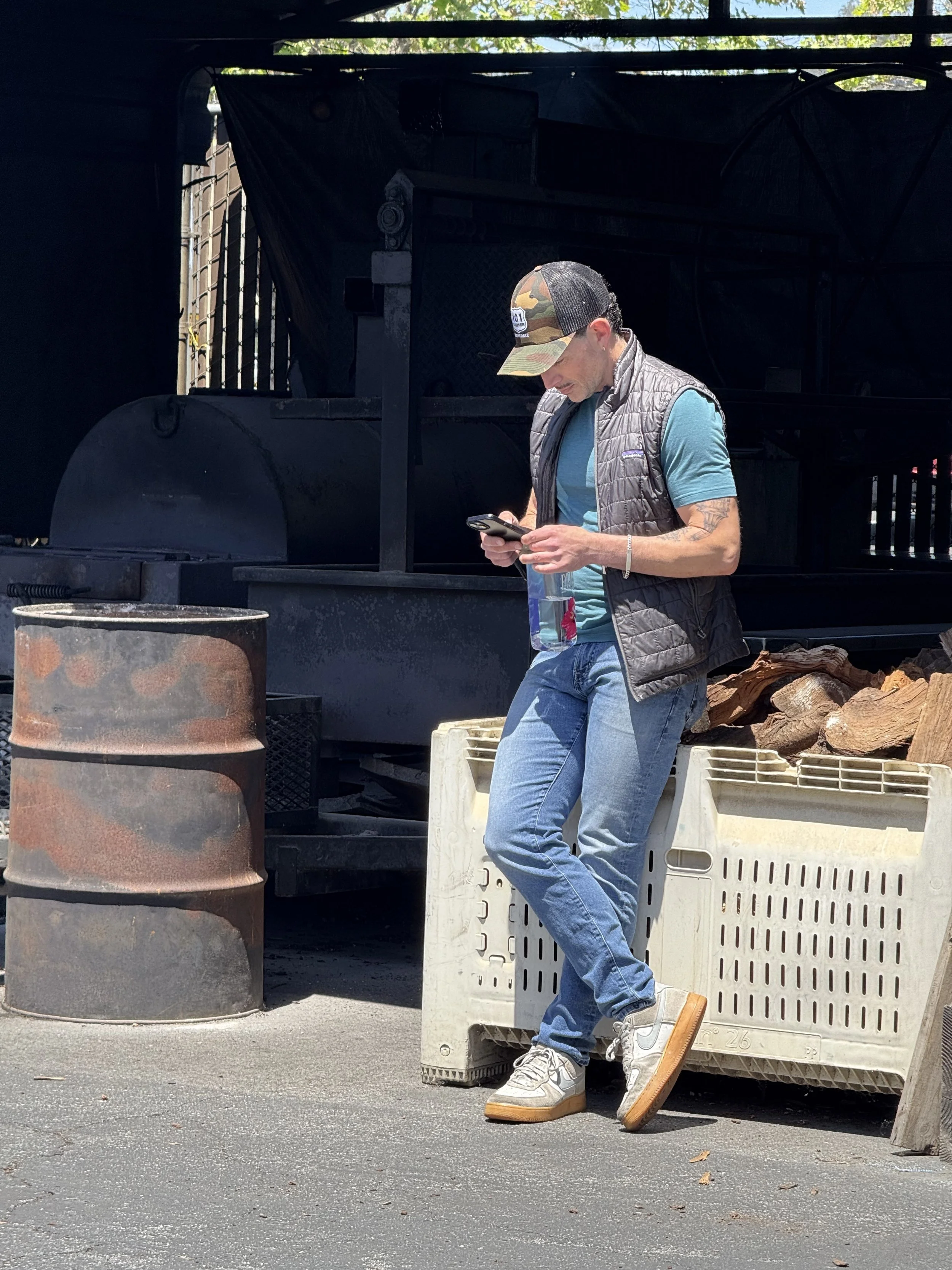 A man standing outdoors, wearing a gray vest, camouflage cap, blue t-shirt, and jeans, looking at his phone, with a plastic bottle in hand and a water bottle in his pocket, leaning against a plastic crate filled with logs.