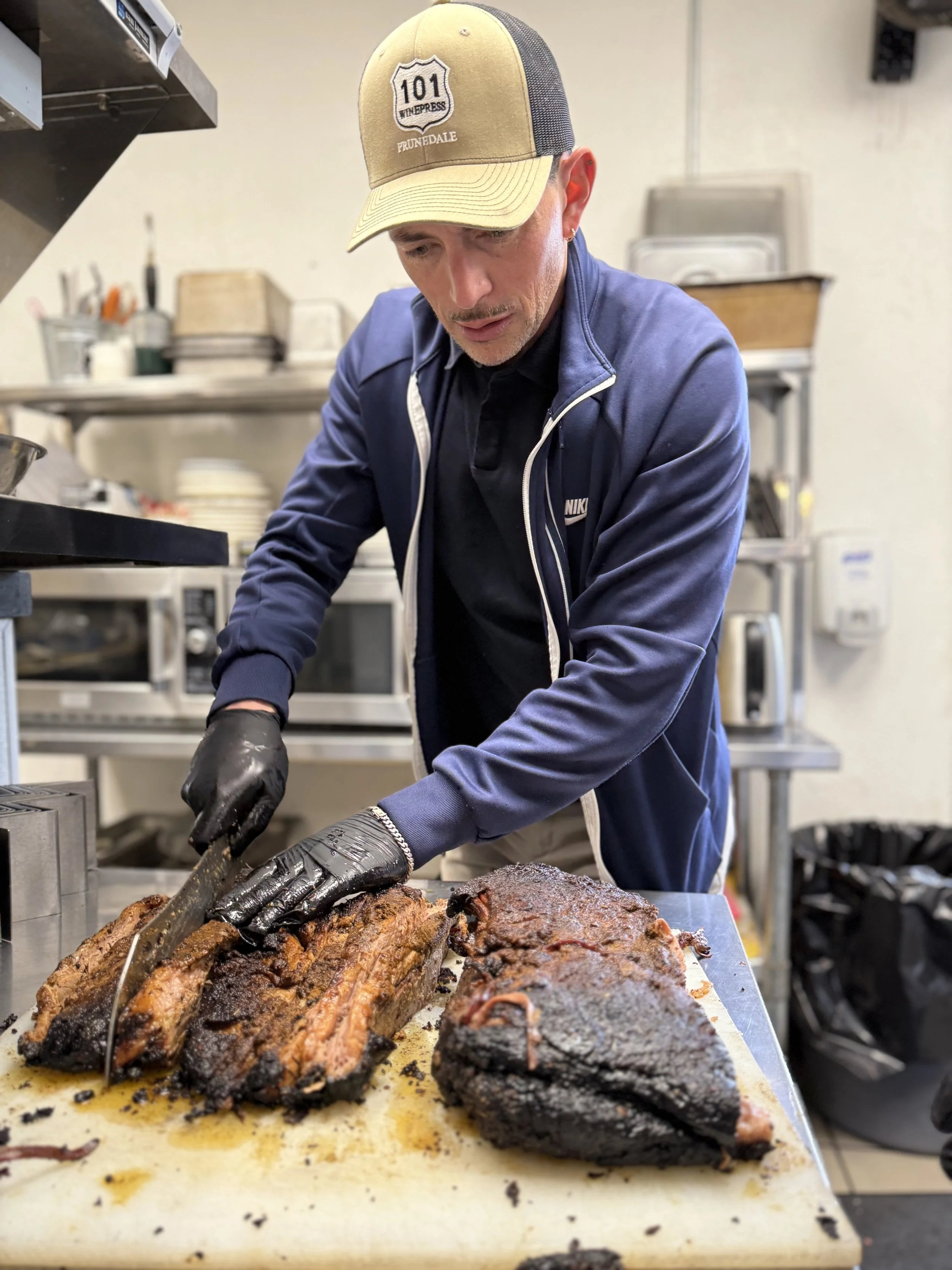 A man with a gray and yellow hat and black gloves slices cooked brisket on a white cutting board in a kitchen.