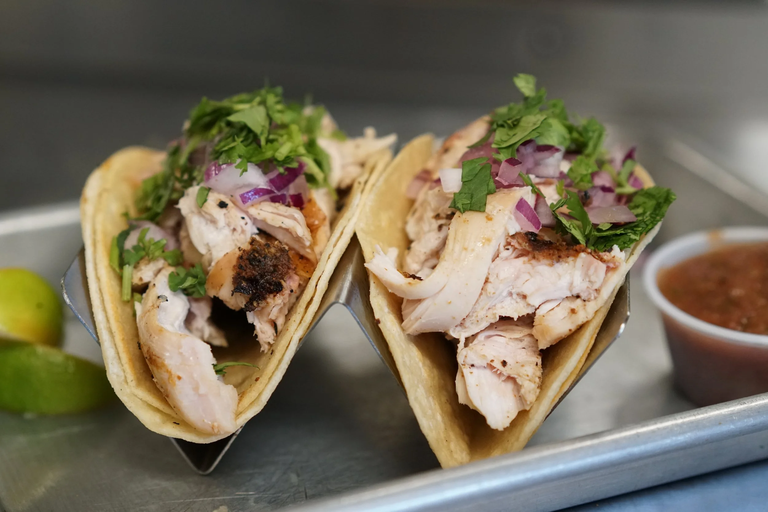 Two fish tacos with grilled fish, chopped onions, cilantro, and a small cup of salsa on a metal tray, with lime wedges on the side.