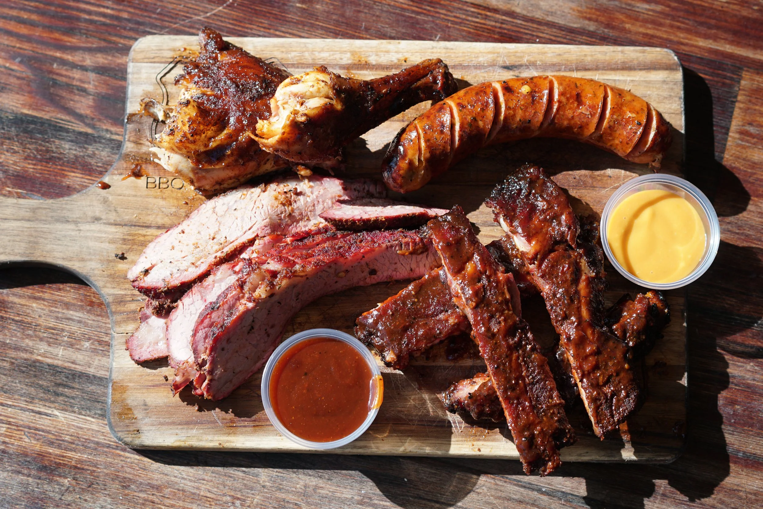 Barbecue platter with sliced brisket, chicken, ribs, sausage, and two dipping sauces on a wooden cutting board.