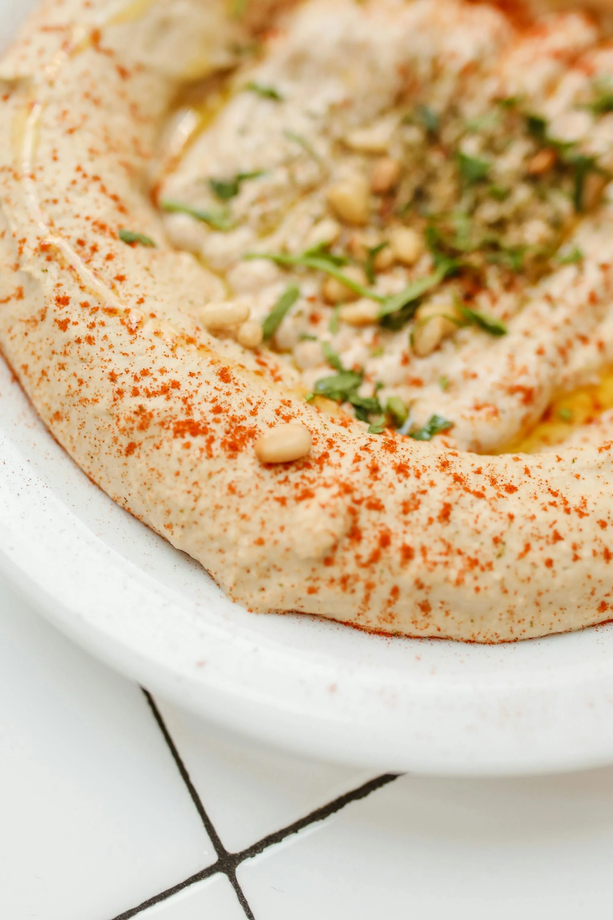 Close-up of a plate of hummus garnished with pine nuts, chopped herbs, and paprika.