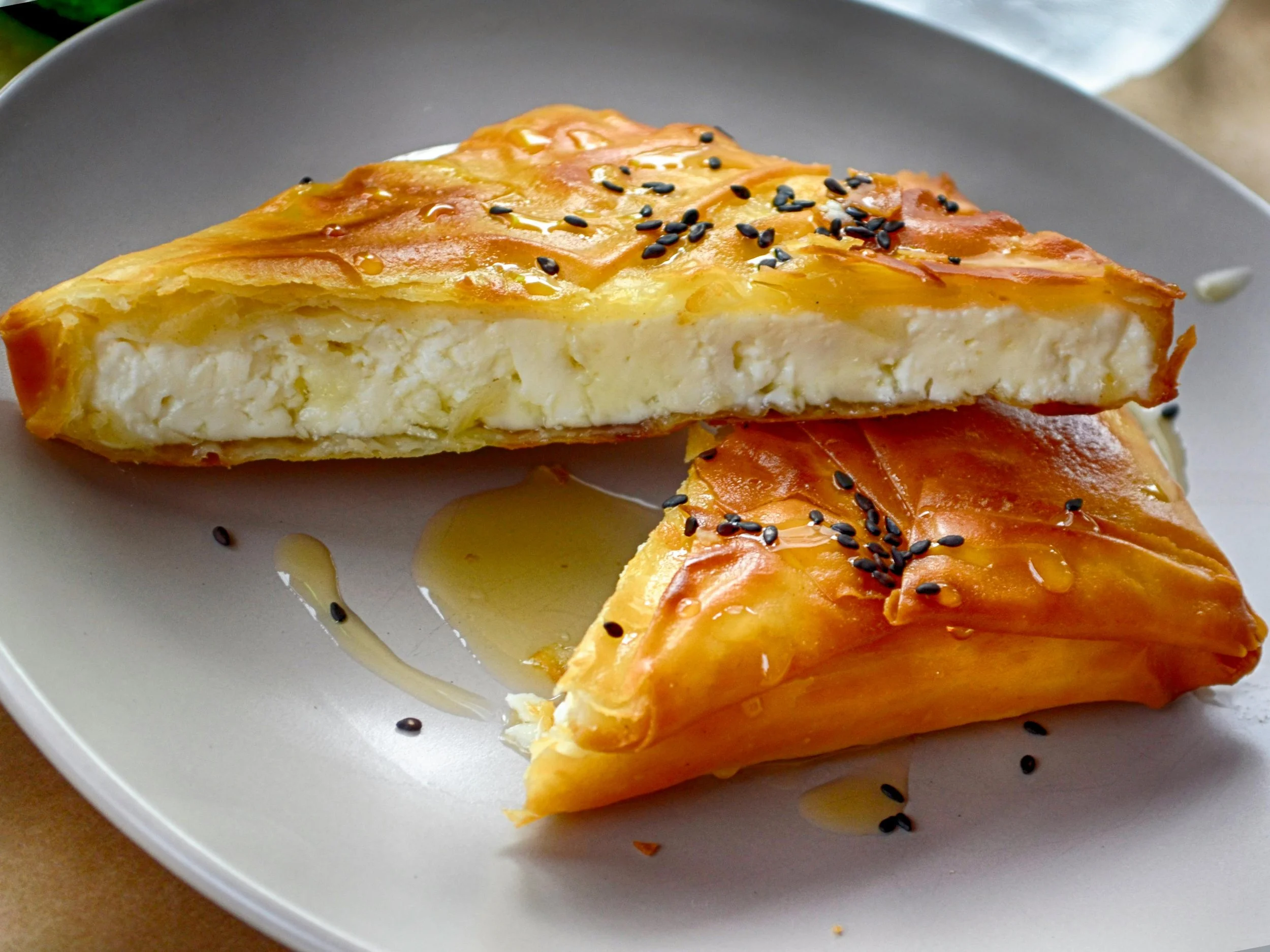 A plate with two pieces of cheesy, flaky pastry sprinkled with black sesame seeds, drizzled with honey.