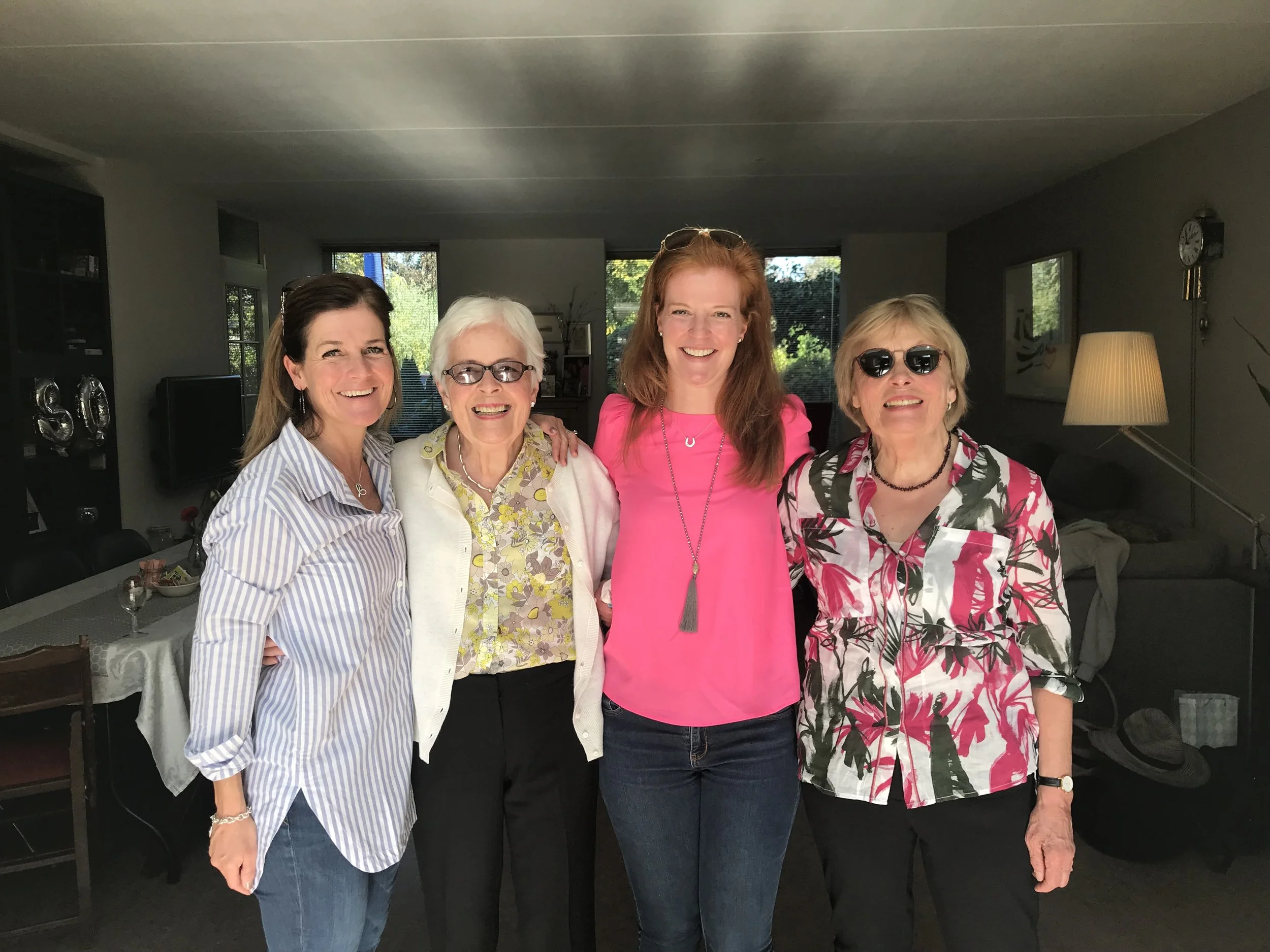Five women standing together inside a living room, smiling at the camera. The room has large windows, a lamp, and a wall clock in the background.