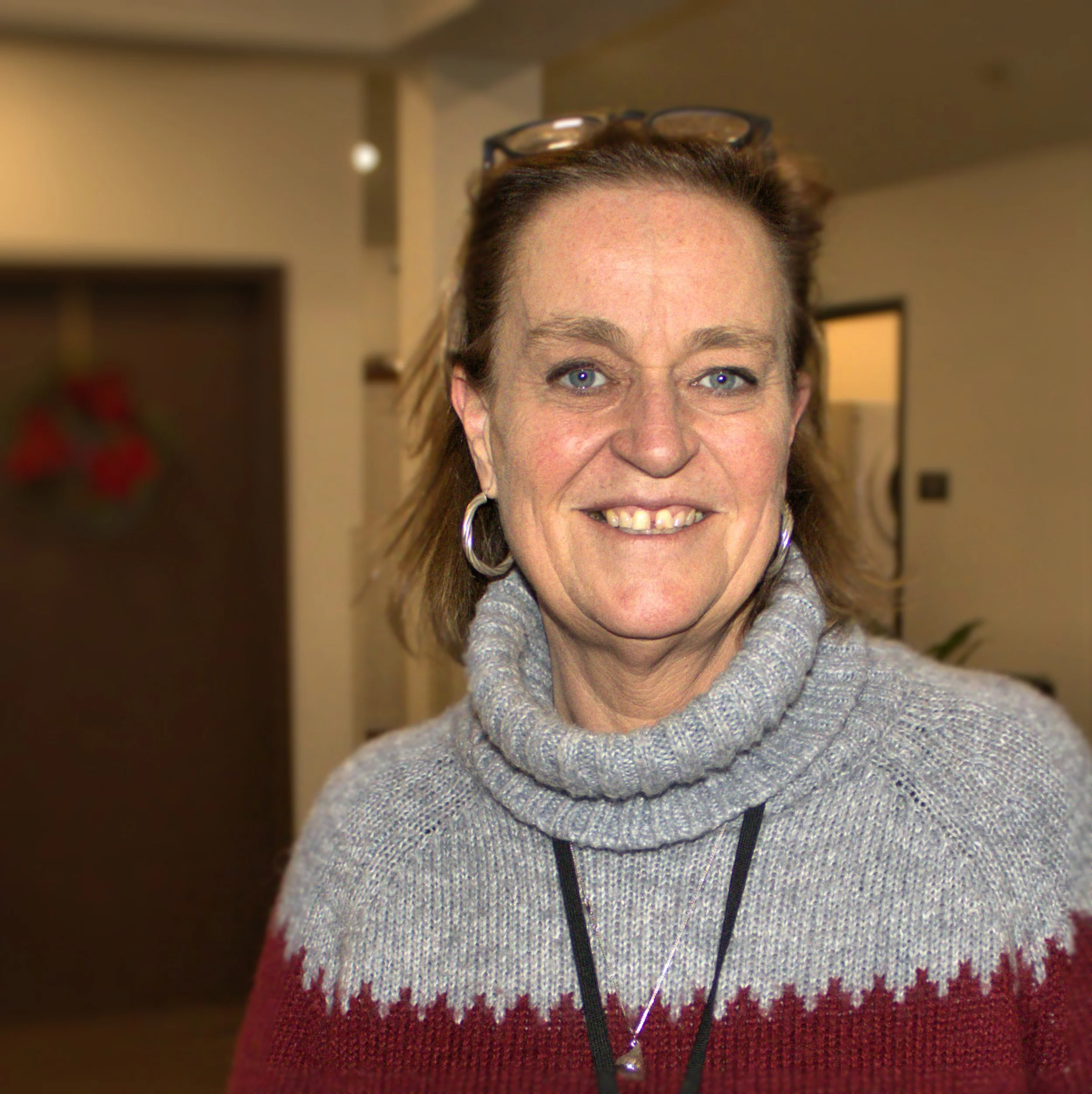 A smiling woman with shoulder-length brown hair, blue eyes, wearing glasses on her head, hoops earrings, and a two-tone gray and red knit sweater, standing indoors.