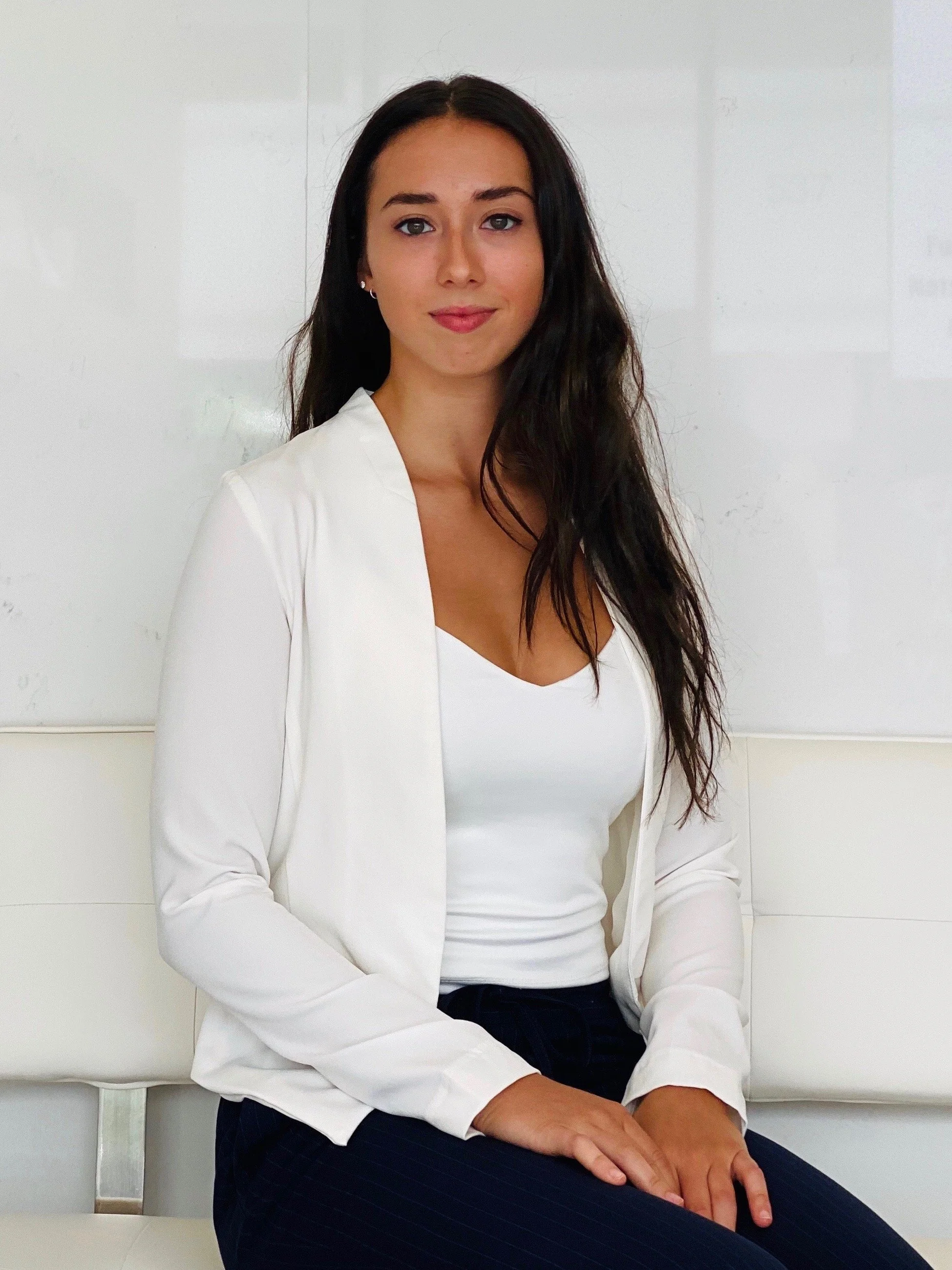 A woman sitting on a white bench, wearing a white shirt and dark pinstriped pants, with dark hair and earrings, against a white background.