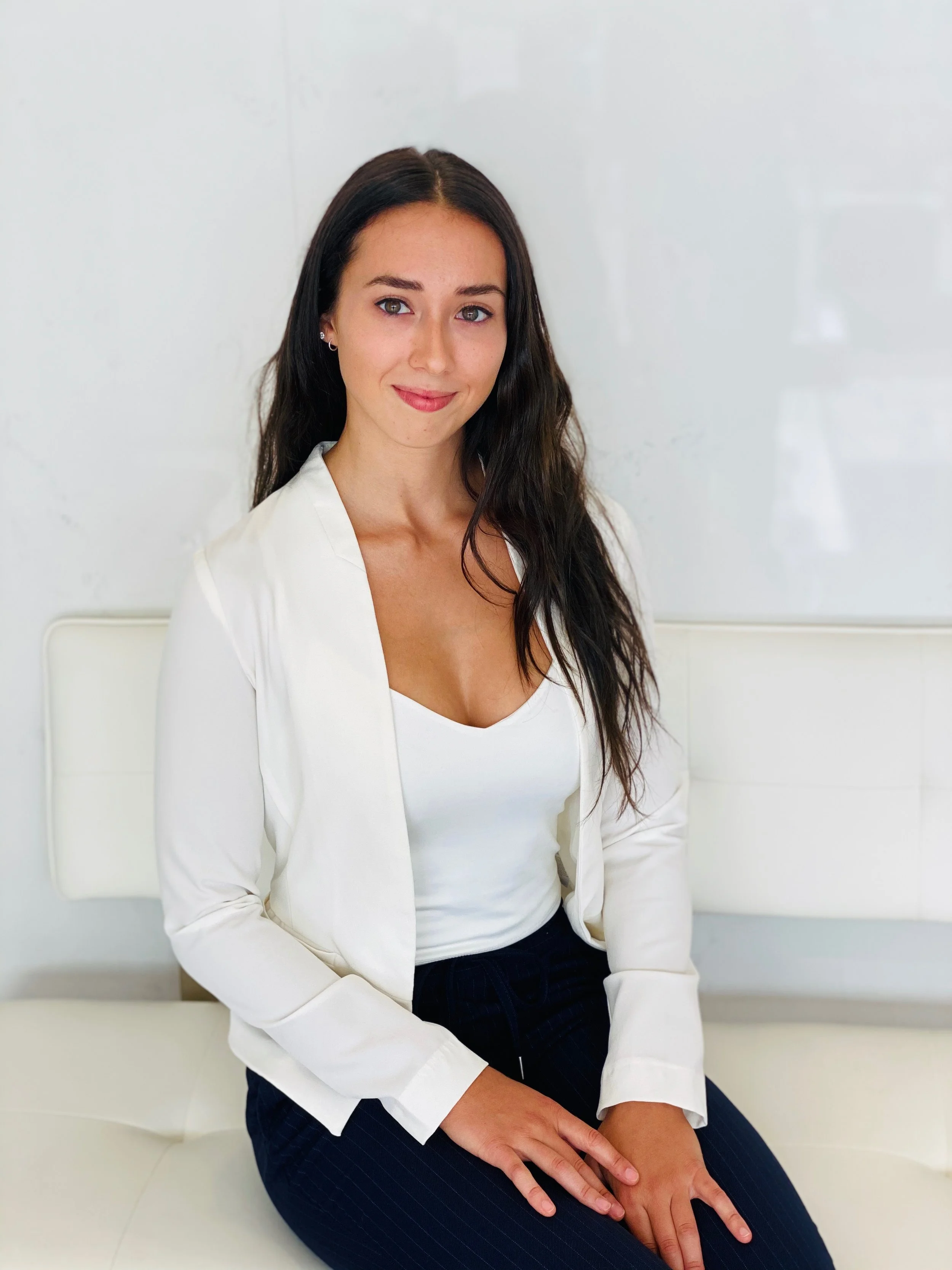 A young woman with long dark hair sitting on a white bench against a plain white wall. She is wearing a white blazer over a white top and dark pants. She is smiling gently and has her hands resting on her lap.