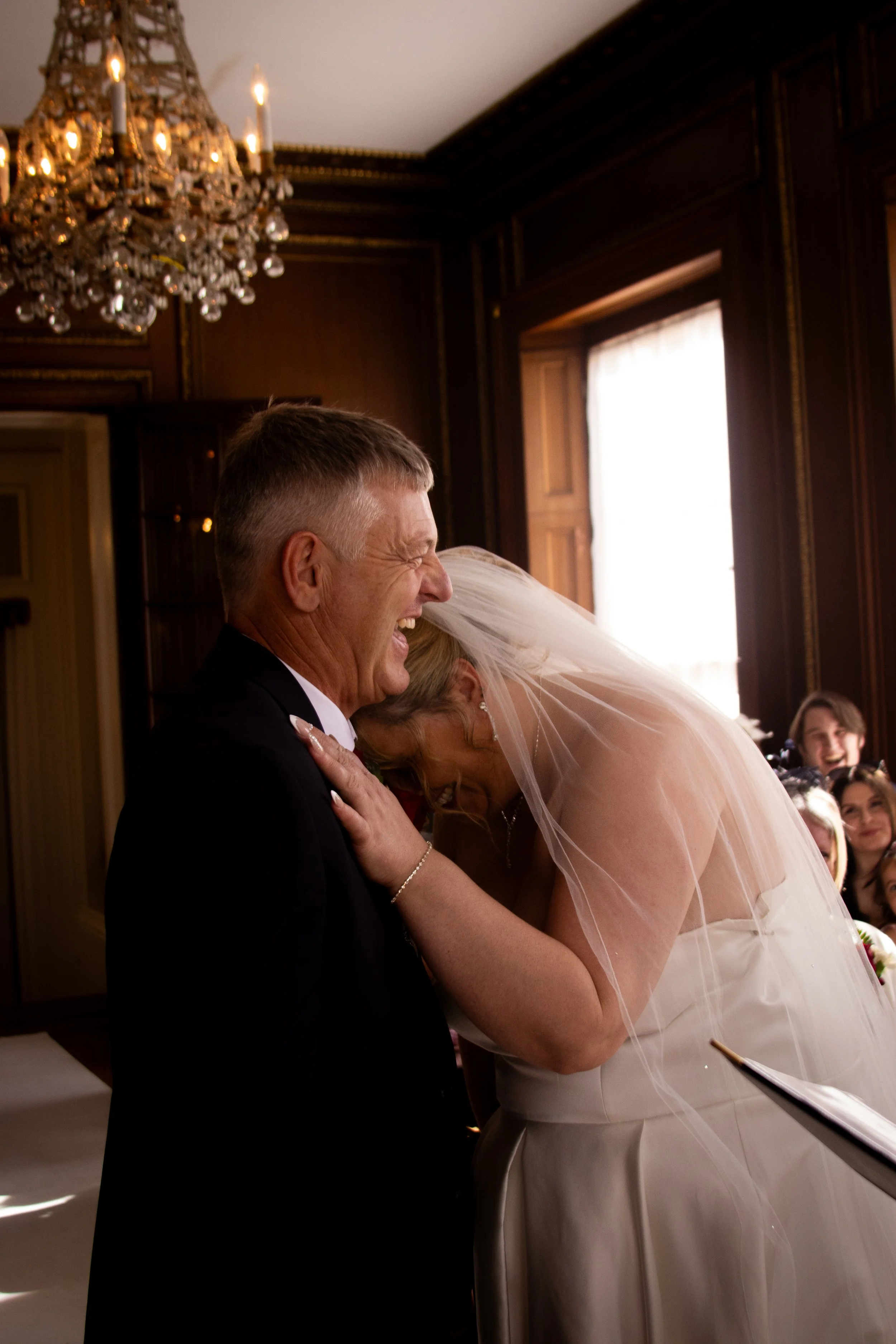 A bride and groom sharing a joyful moment, with the bride leaning on the groom's chest, in a richly decorated room with a chandelier and wooden paneling.