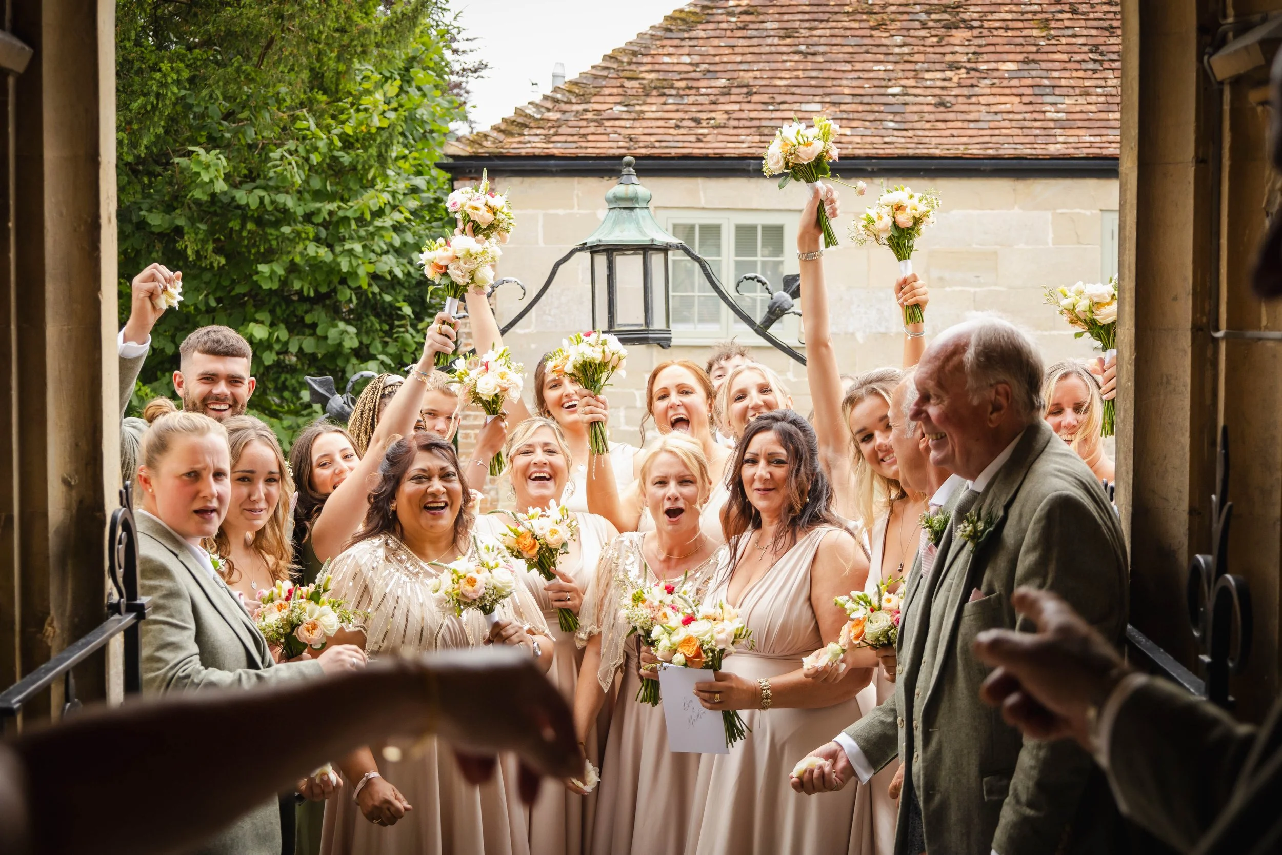 Group of people dressed in wedding attire celebrating and holding bouquets of flowers outdoors.