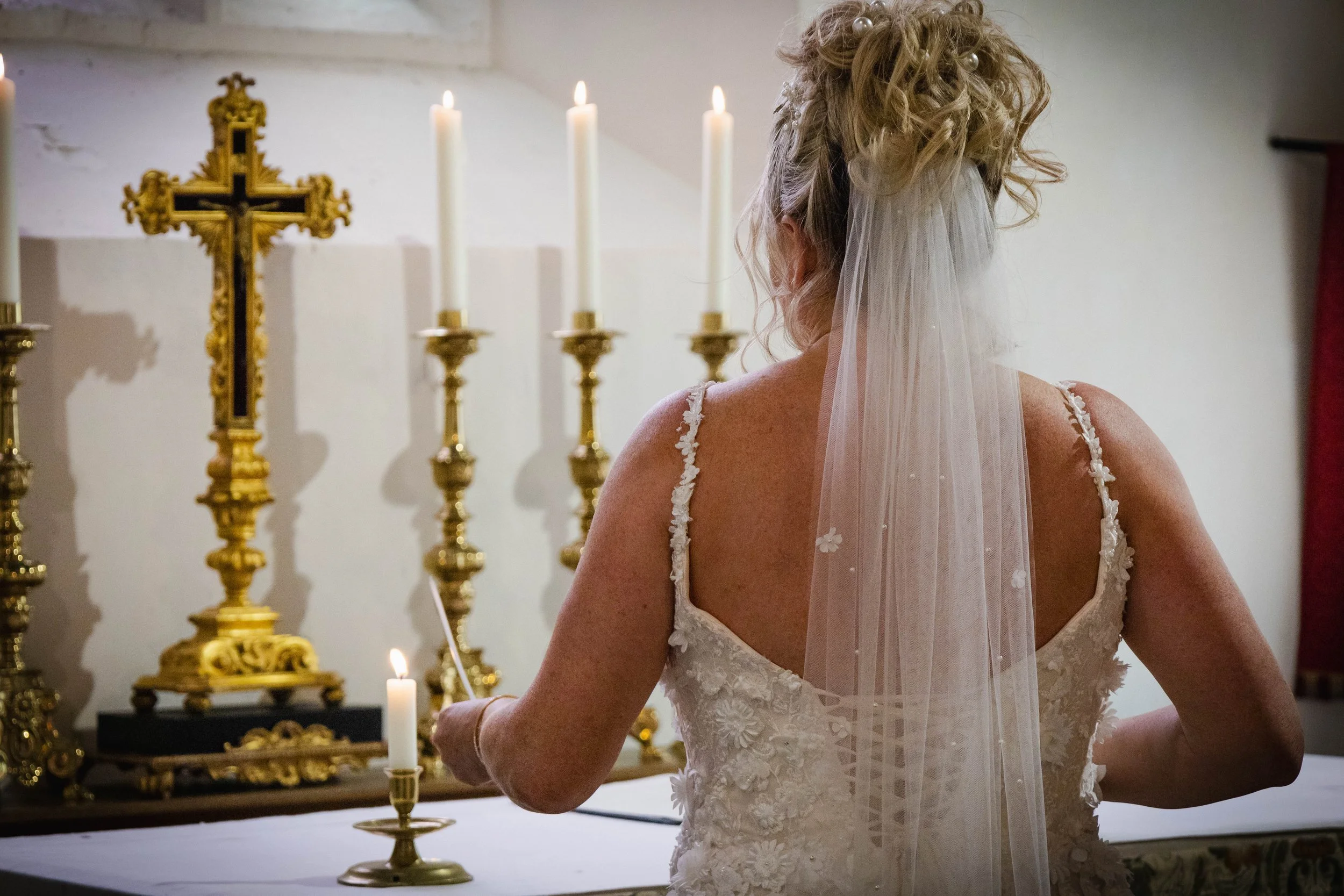 A bride with curly blonde hair in a bridal gown and veil standing in front of an altar with lit candles and a gold cross in a church.