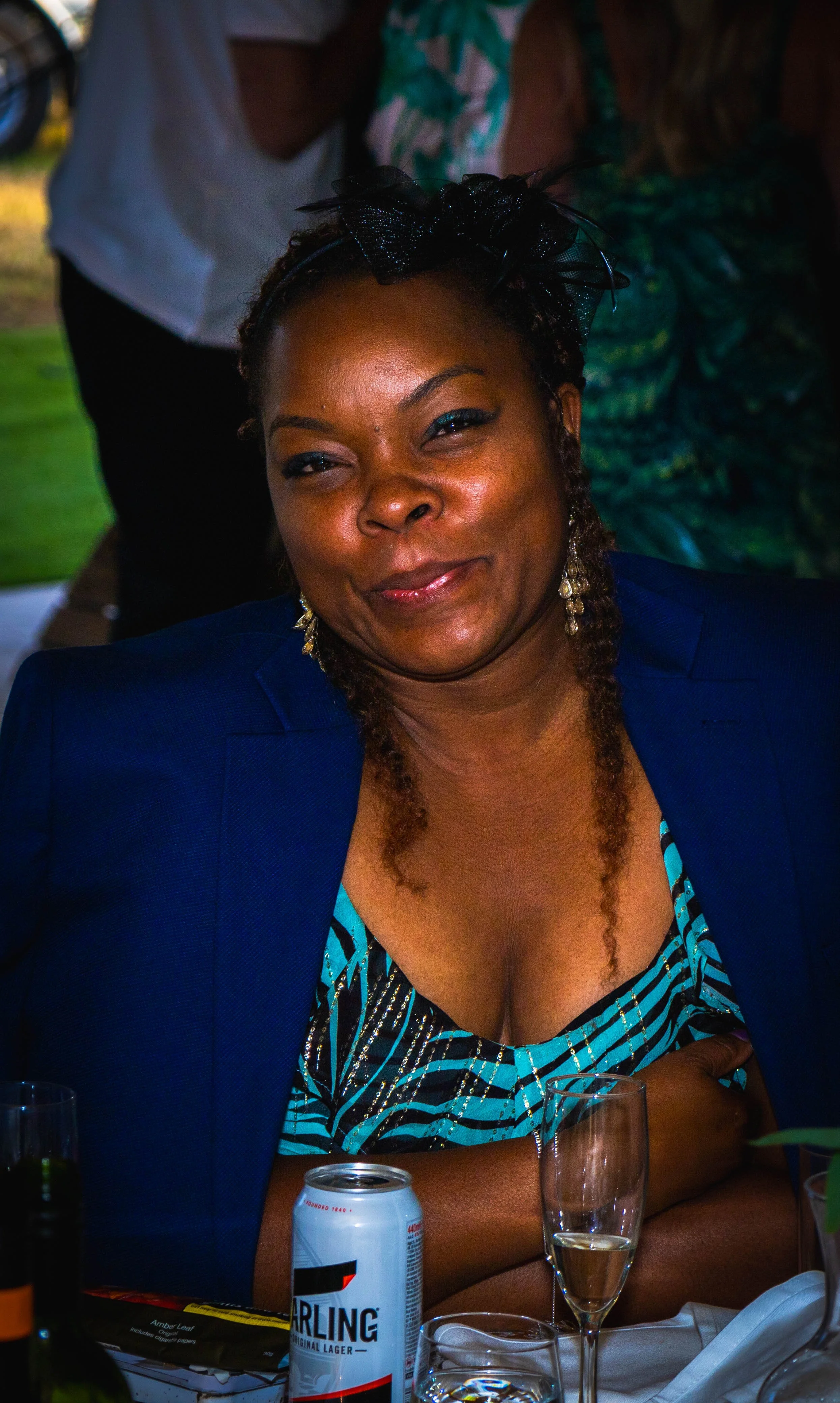 A woman with braided hair, wearing a blue blazer and patterned top, sitting at a table with a can of Sparkling beer, a glass of champagne, and some food items, at an indoor or outdoor event.